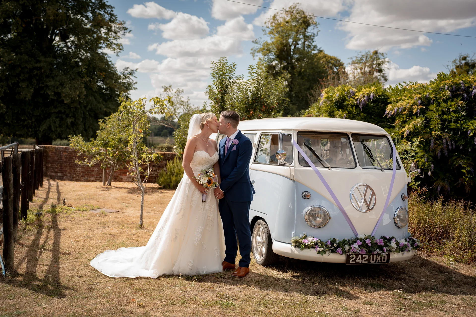 A newlywed couple shares a tender kiss next to a pale blue and white vintage VW campervan decorated with ribbons and floral displays outdoors on a sunny summer day.
