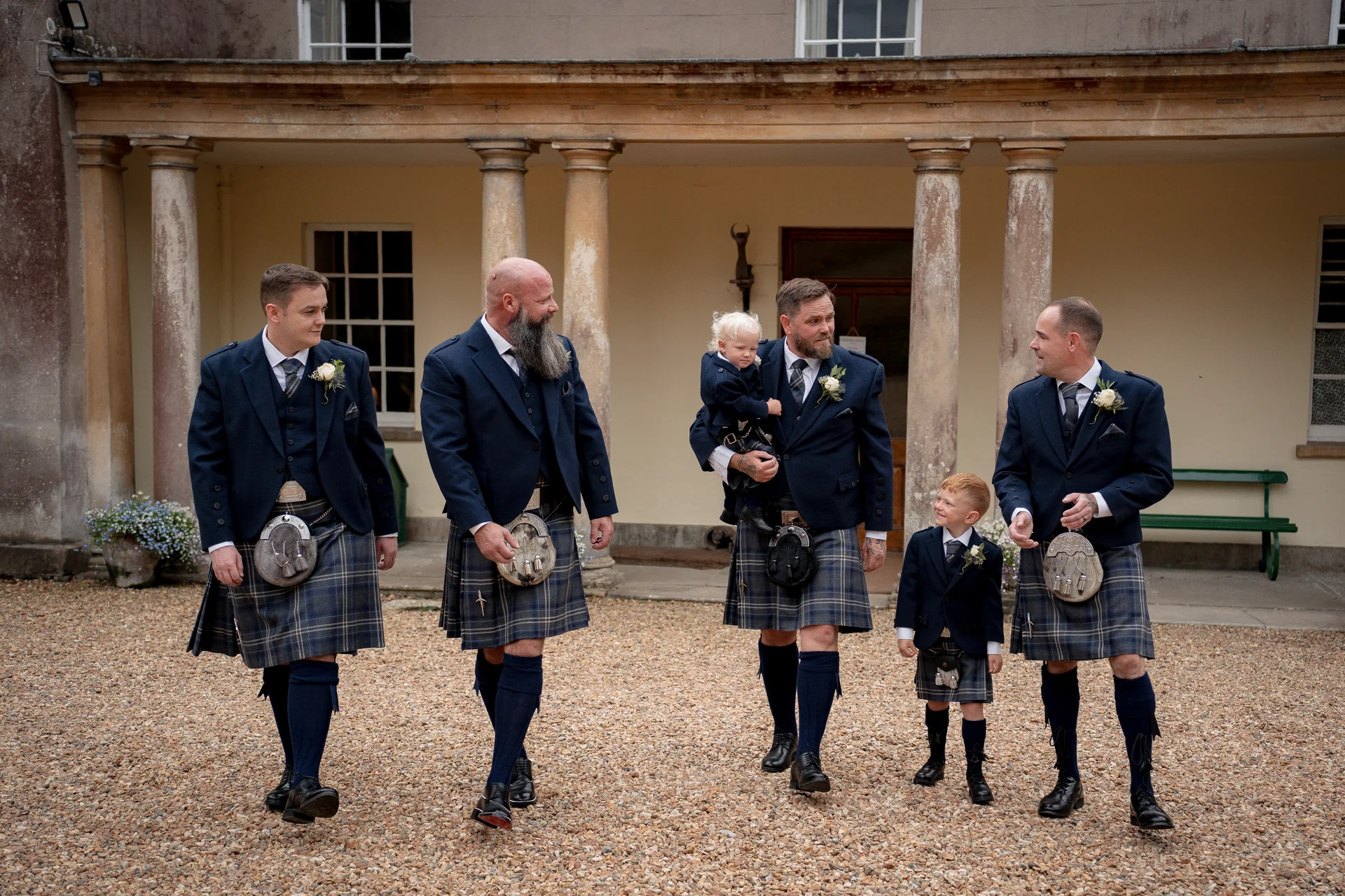 A group of men and a young boy dressed in traditional Scottish kilts walk together across a gravel courtyard in front of a grand manor house.