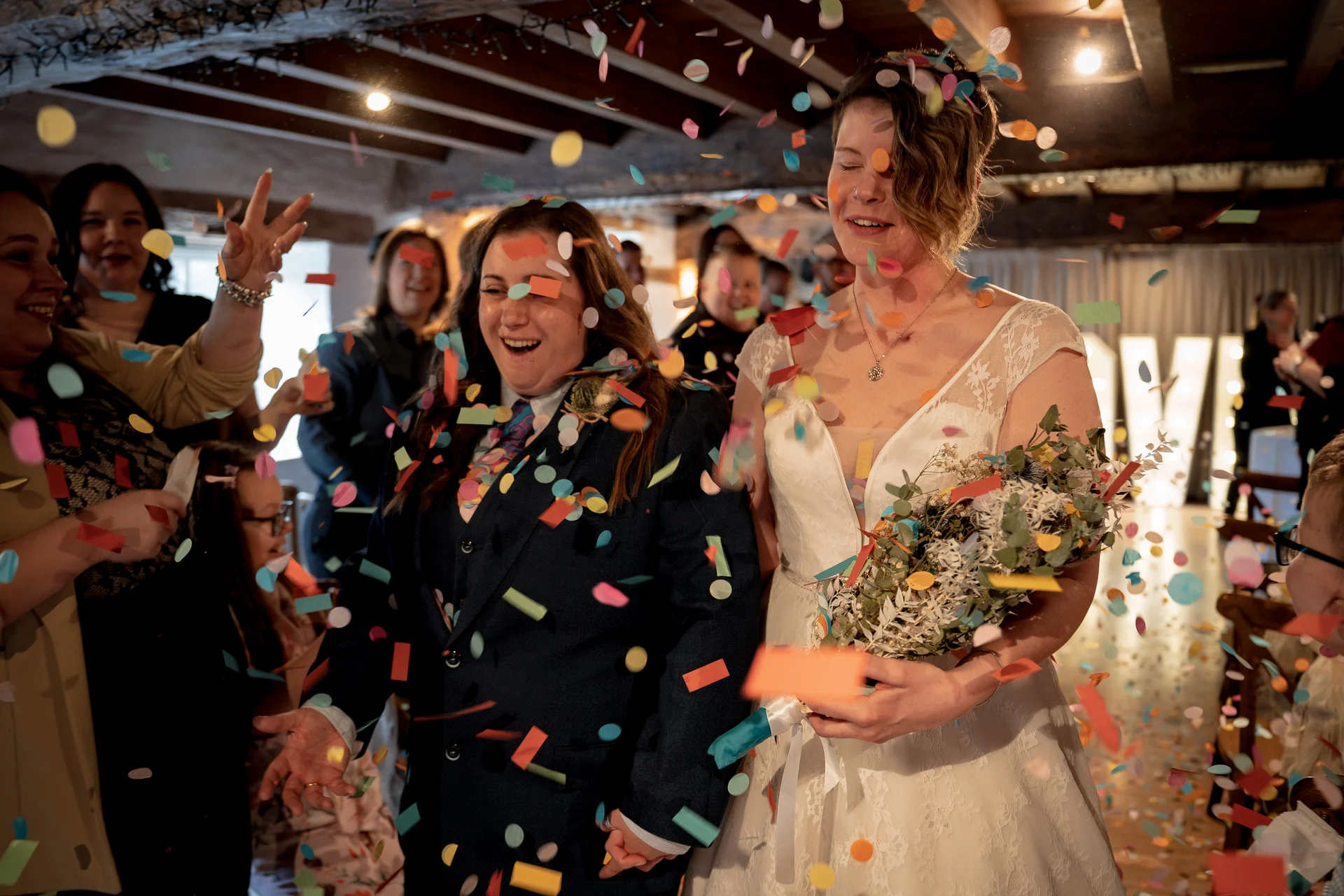 Two smiling brides walk through a shower of colourful confetti inside a rustic barn venue with exposed wooden beams.