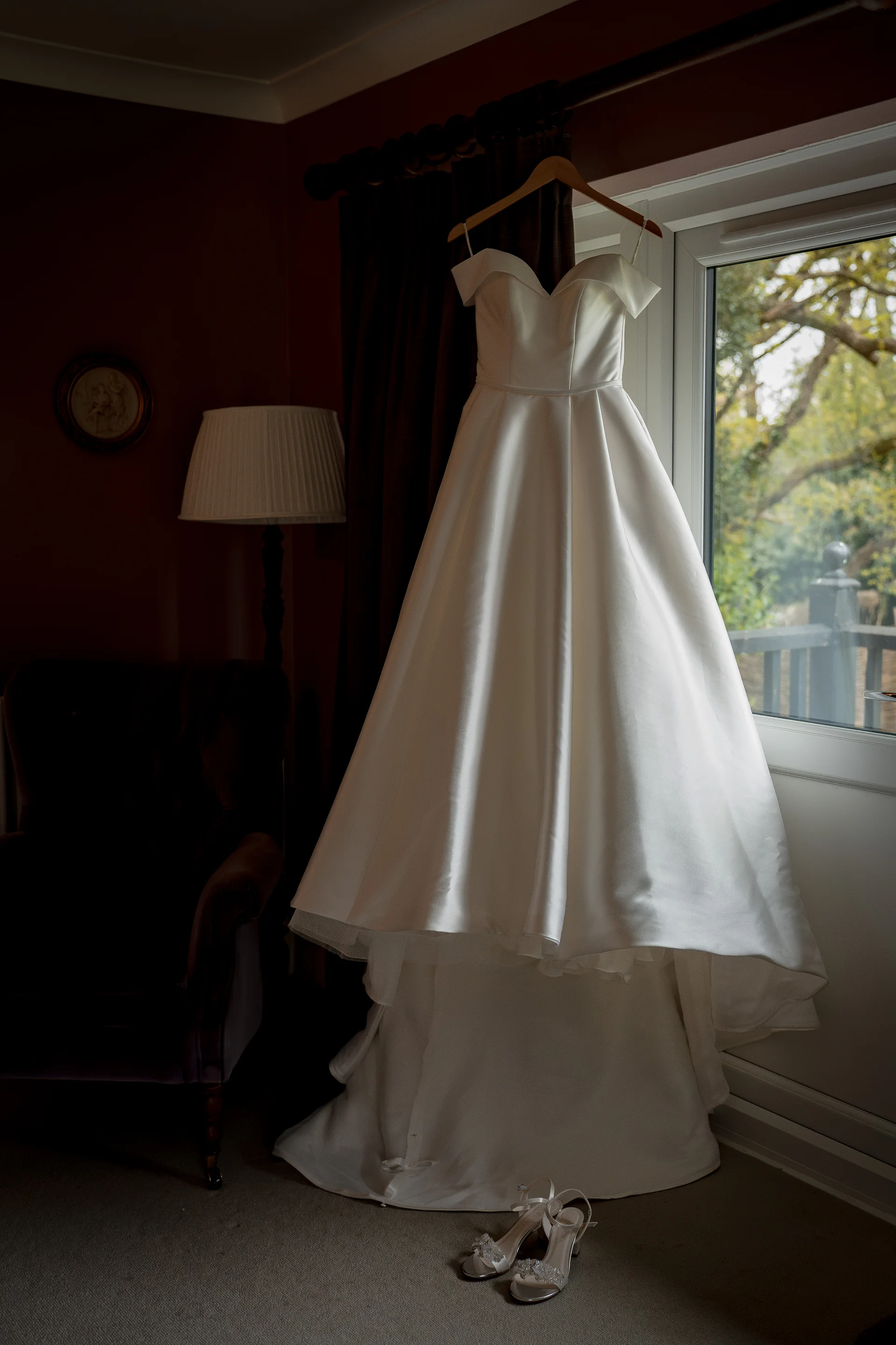 An off-the-shoulder satin a-line wedding dress hangs by a bright window, with matching embellished bridal heels resting on the floor below.