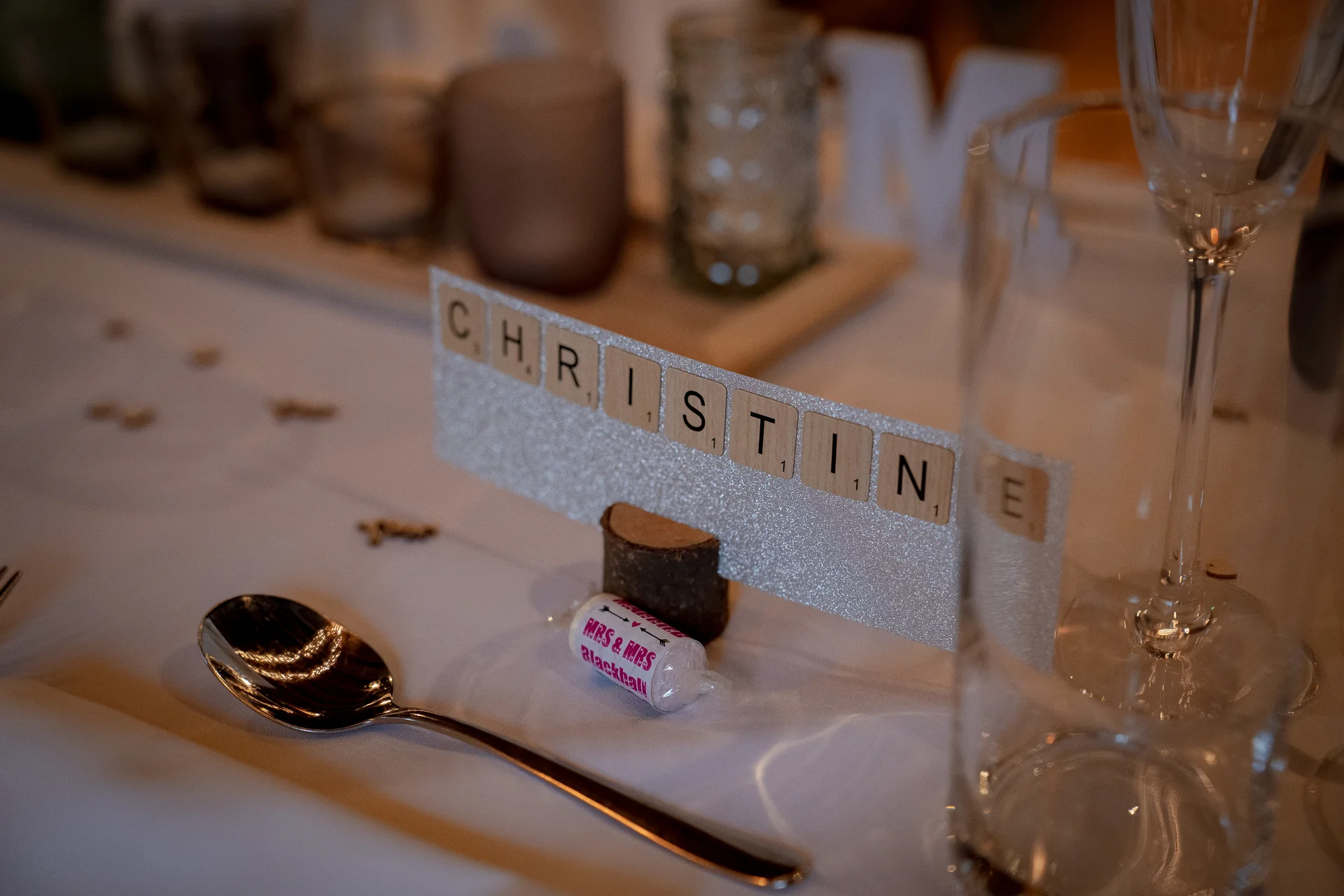 A rustic place setting features a glittering name card spelled out in Scrabble tiles next to a polished spoon and crystal glassware.