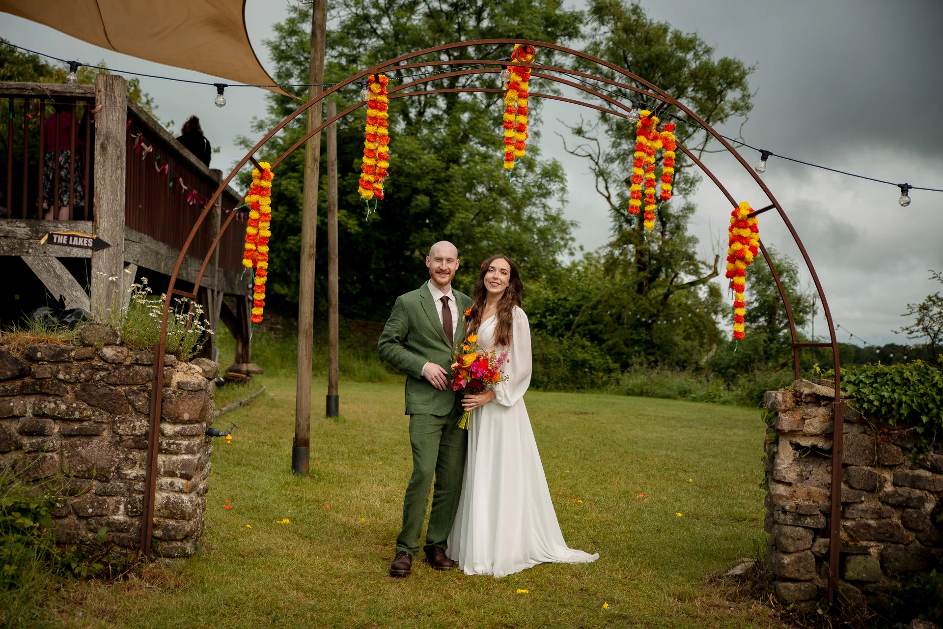A bride in a white long-sleeve gown and groom in a green suit stand smiling together beneath a metal ceremony arch adorned with vibrant marigold floral garlands outdoors.
