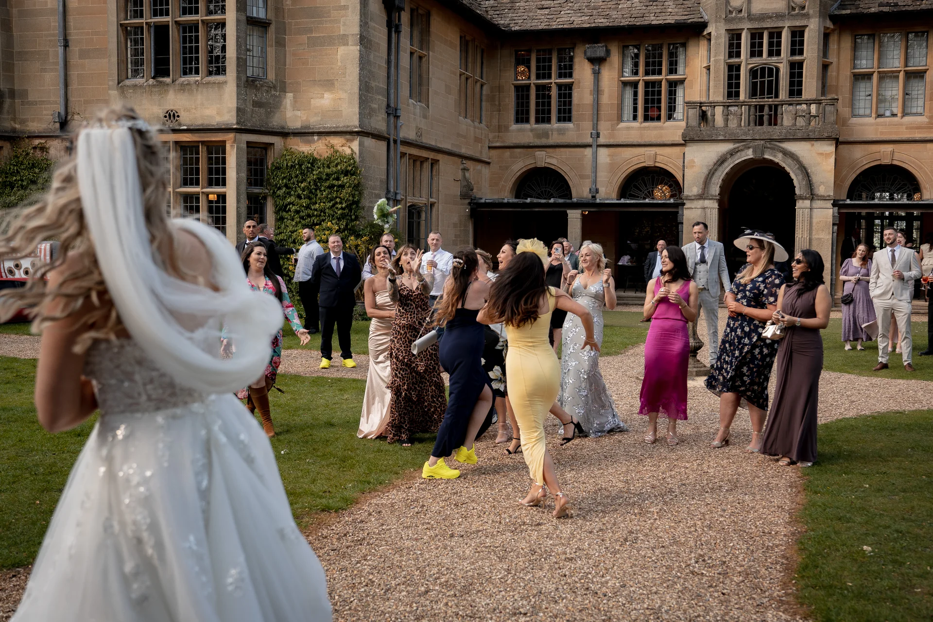 A bride in a white wedding gown prepares to throw her bouquet to a group of excited wedding guests gathered in the gravel courtyard of a grand stone manor house.