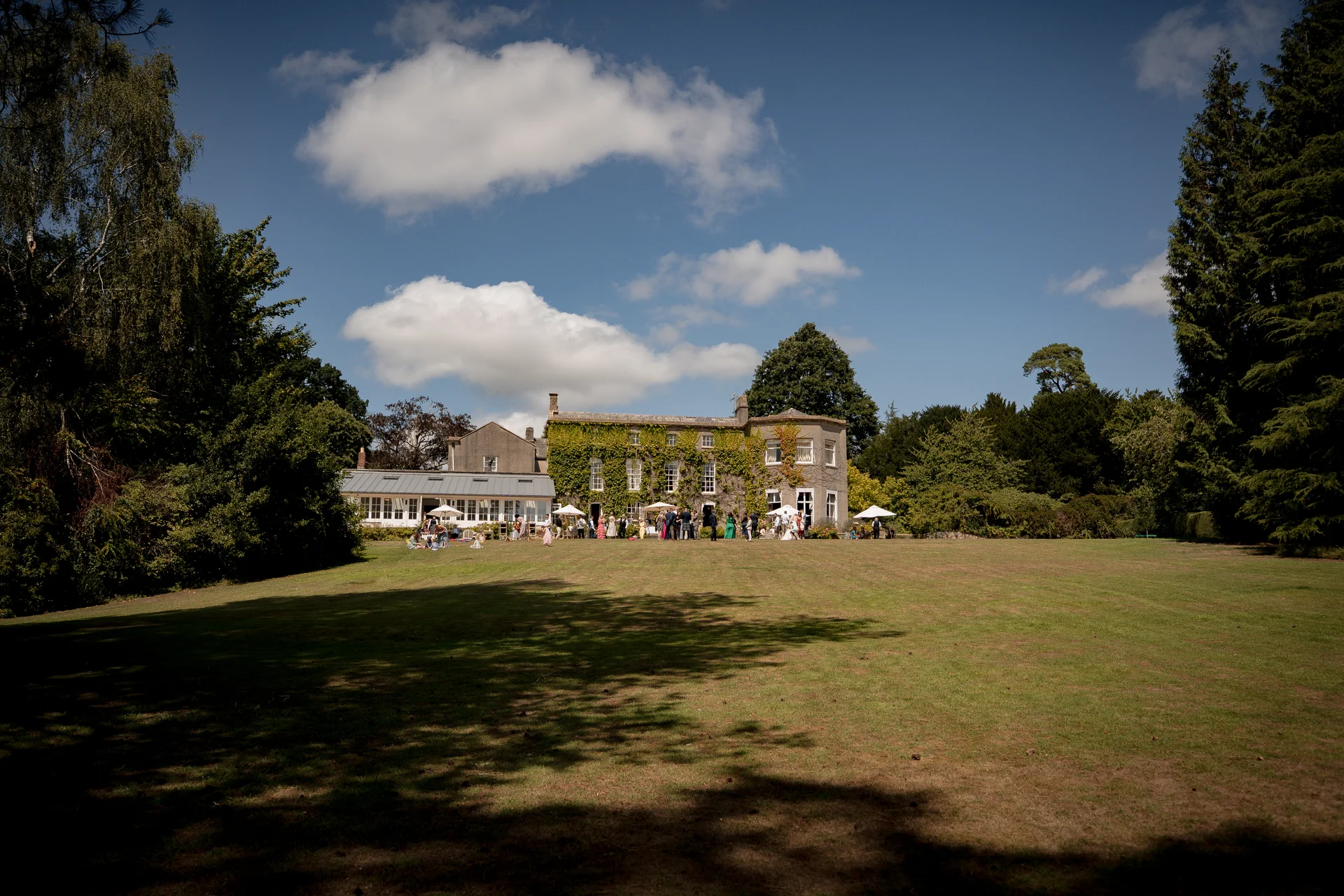 Guests enjoy a sunny drinks reception on the sprawling lawn in front of an ivy-clad country manor house under a bright blue sky.
