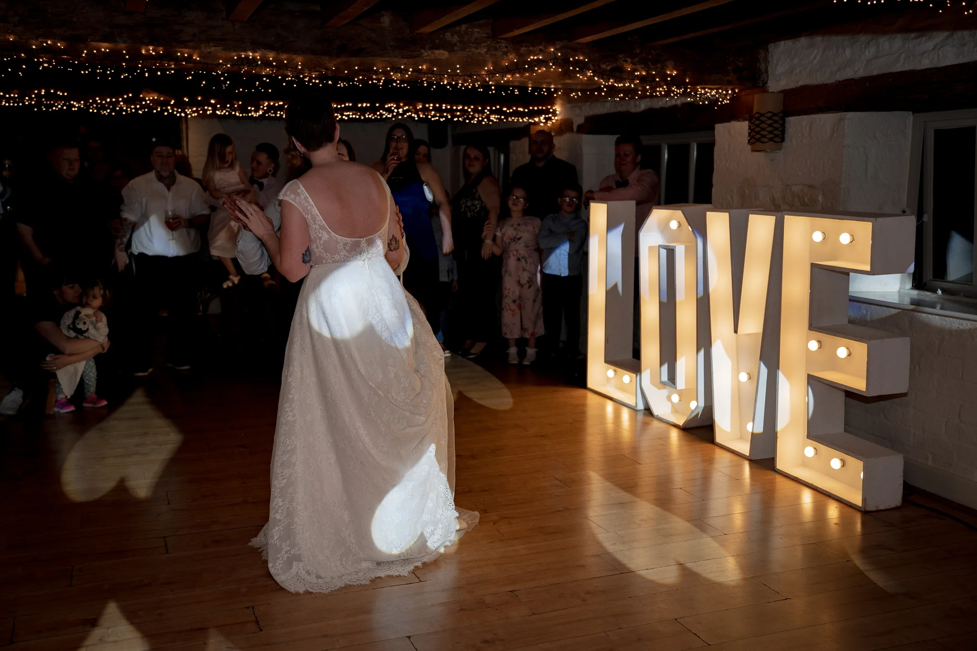 A newlywed couple shares their first dance on a wooden floor in a rustic barn venue illuminated by fairy lights and giant light-up love letters.