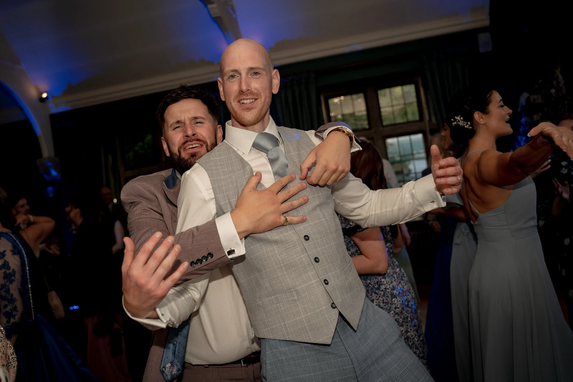 Two smiling groomsmen embrace playfully on a lively evening wedding dance floor illuminated by blue ambient party lighting.