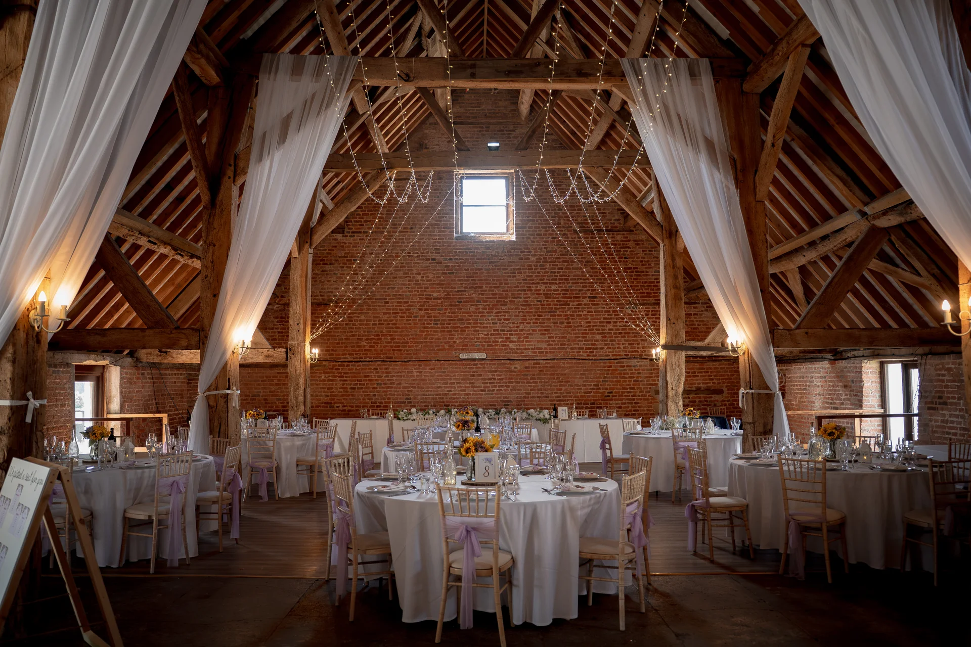 A rustic barn wedding reception featuring exposed brick walls, wooden beams, a fairy light canopy, and round tables draped in white linen.