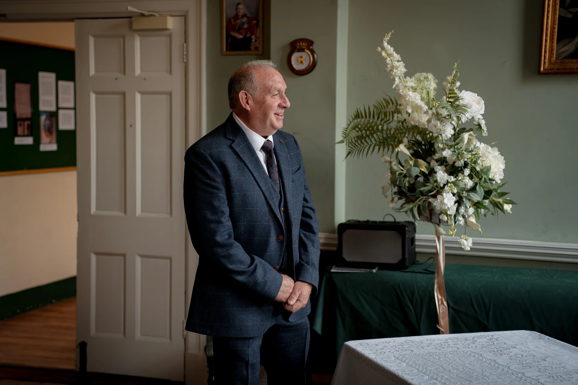 A smiling groom in a checked suit stands inside an interior room next to a large white floral pedestal arrangement.