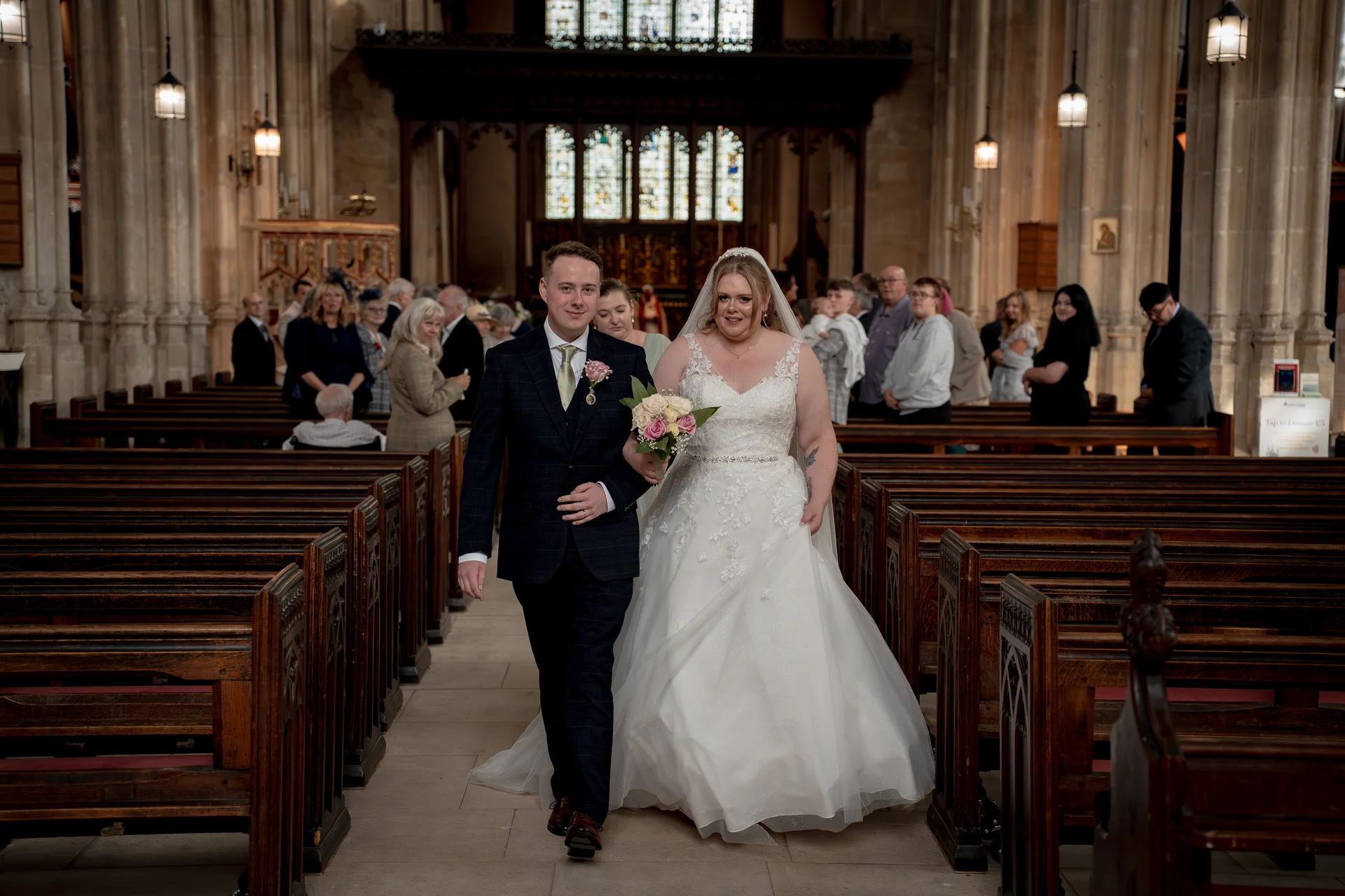 Newlywed bride and groom walk hand in hand down the central aisle of a grand historic church, flanked by wooden pews and watching wedding guests.