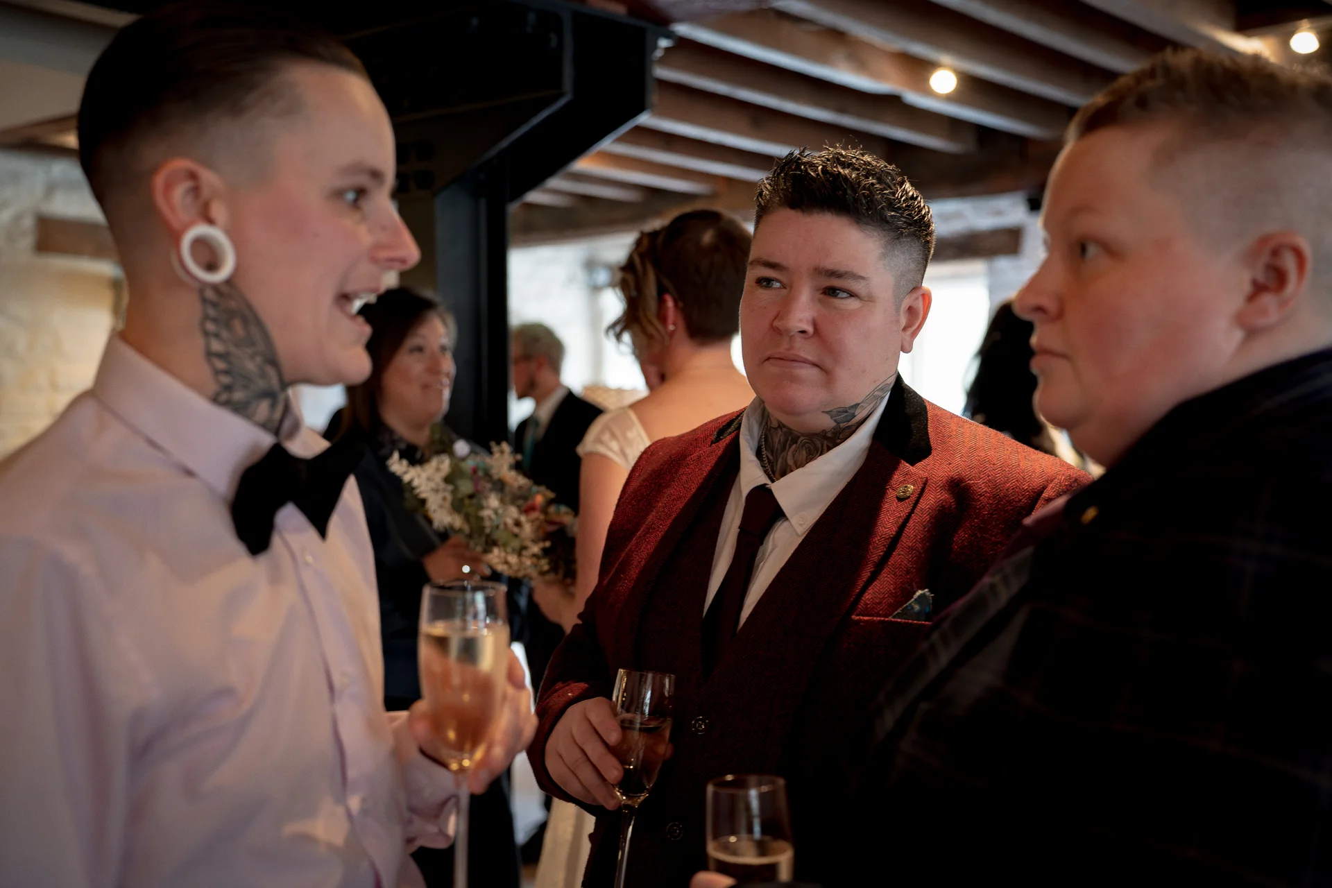 A group of stylishly dressed guests chat and hold glasses of champagne during a lively wedding reception indoors.