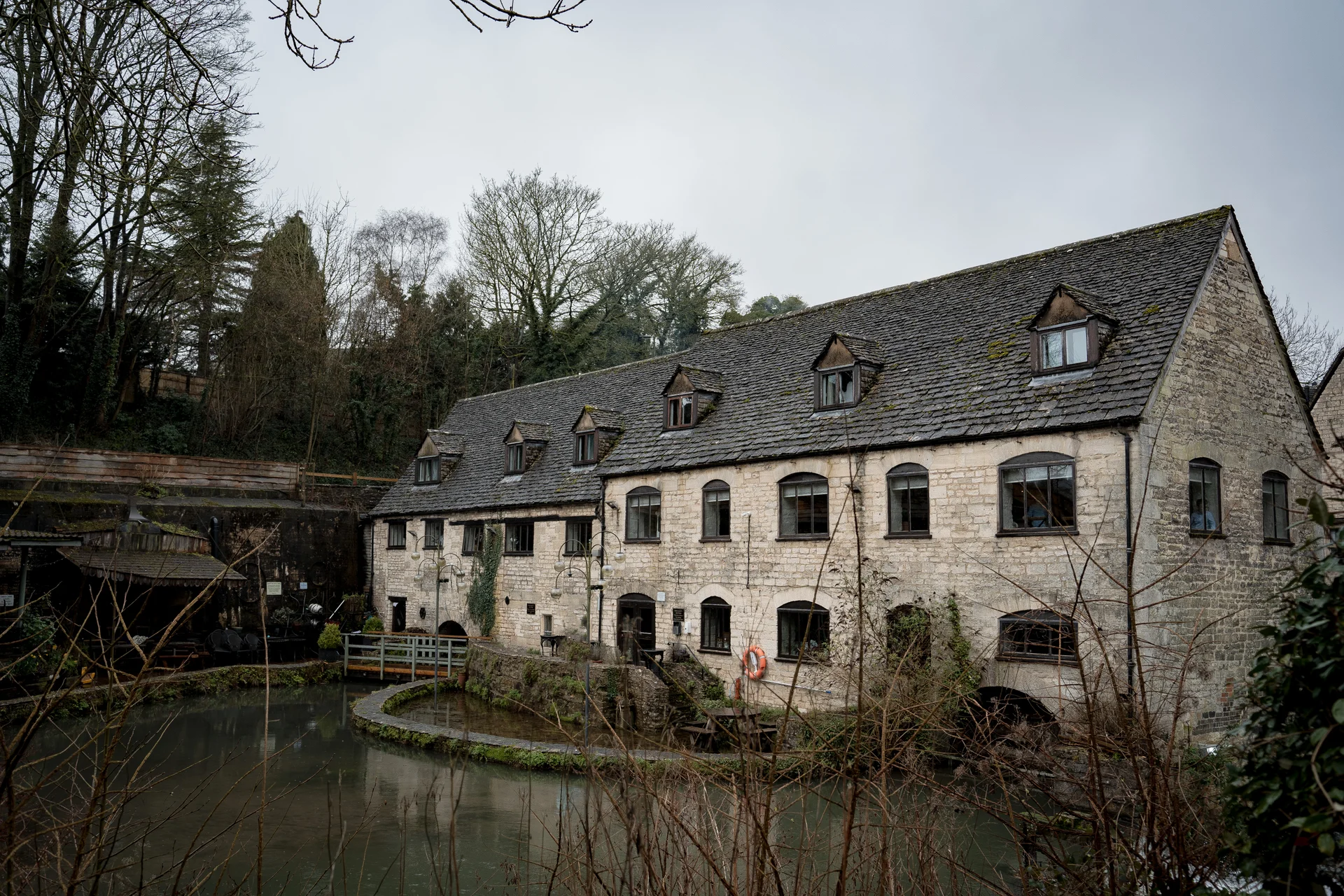A wide exterior shot of a historic stone mill building with dark tiled roof, situated alongside a still pond under an overcast grey sky.