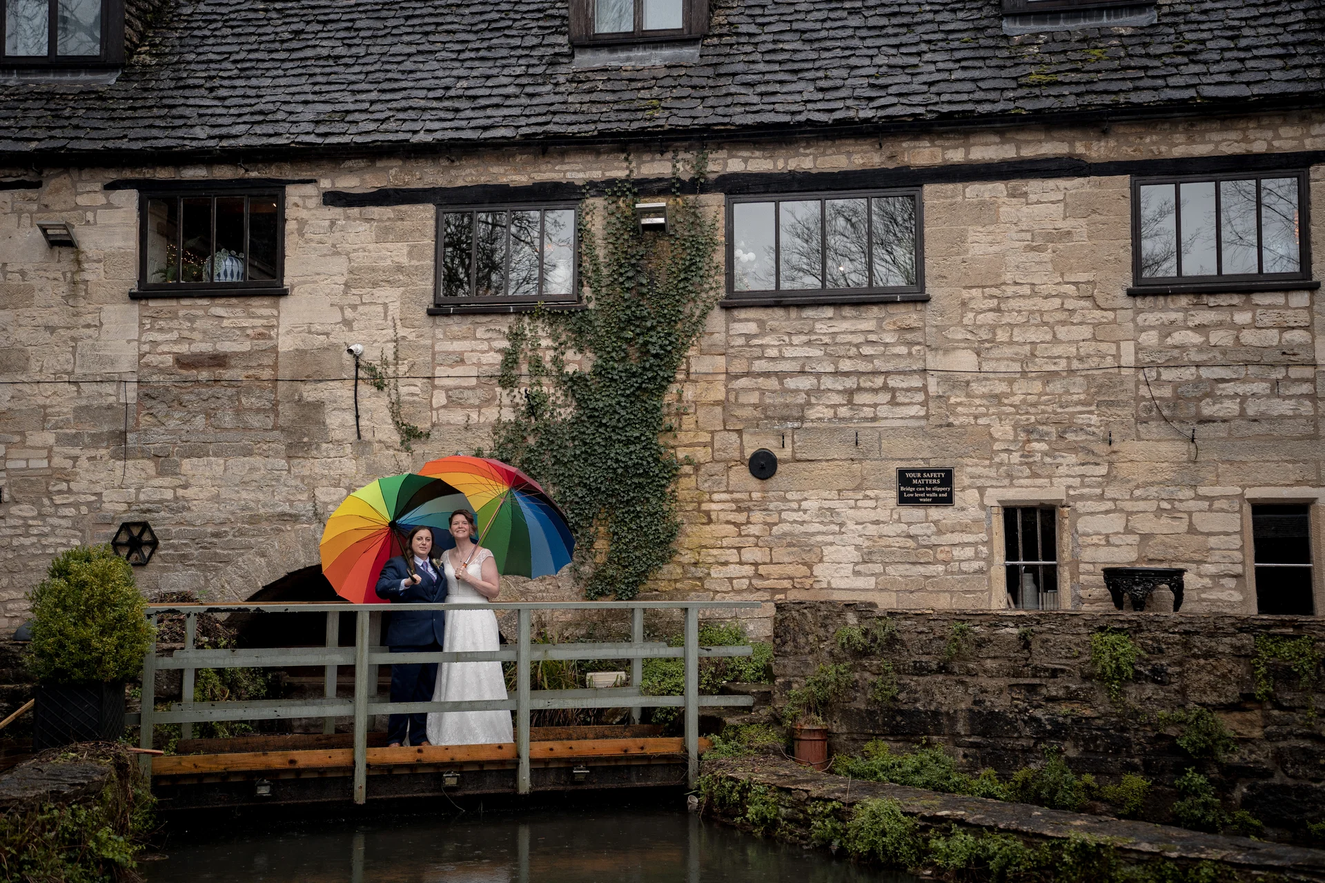 A newlywed same-sex couple stands under a vibrant rainbow umbrella on a wooden bridge in front of a rustic stone building during a rainy wedding day.