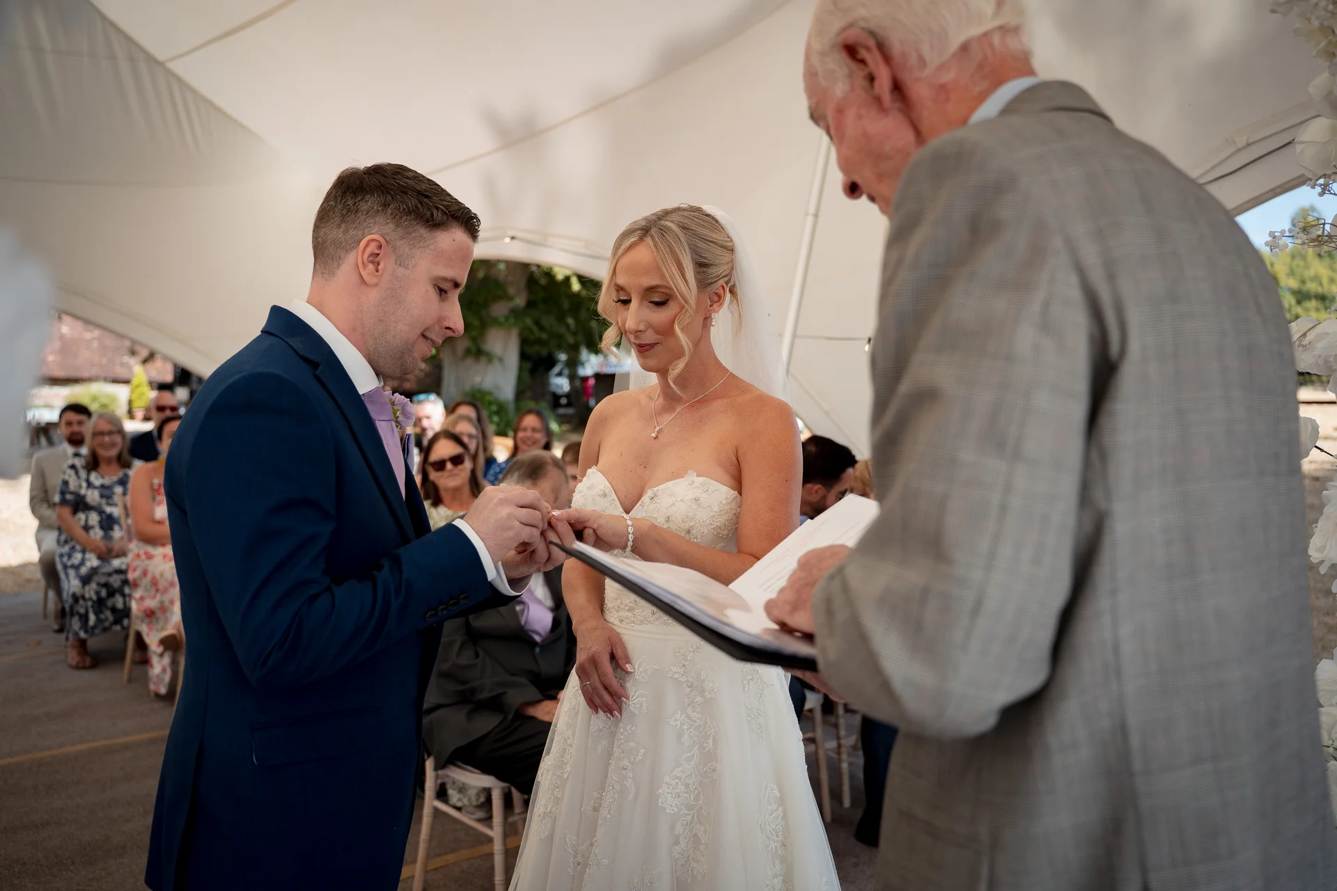 The groom gently places a ring on the bride's finger during their outdoor marquee ceremony, watched closely by the celebrant.