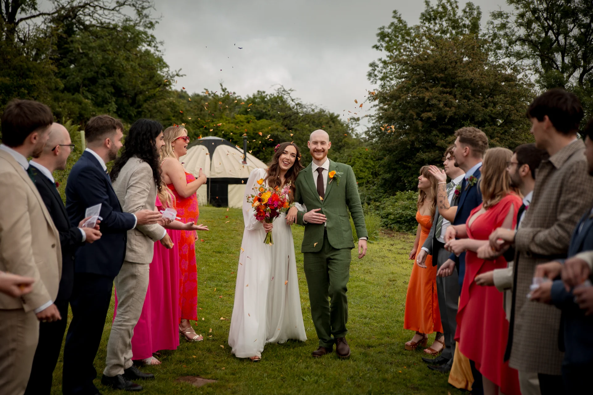 Newlyweds walk hand-in-hand through a grassy field past cheering wedding guests who throw colorful confetti in the air.