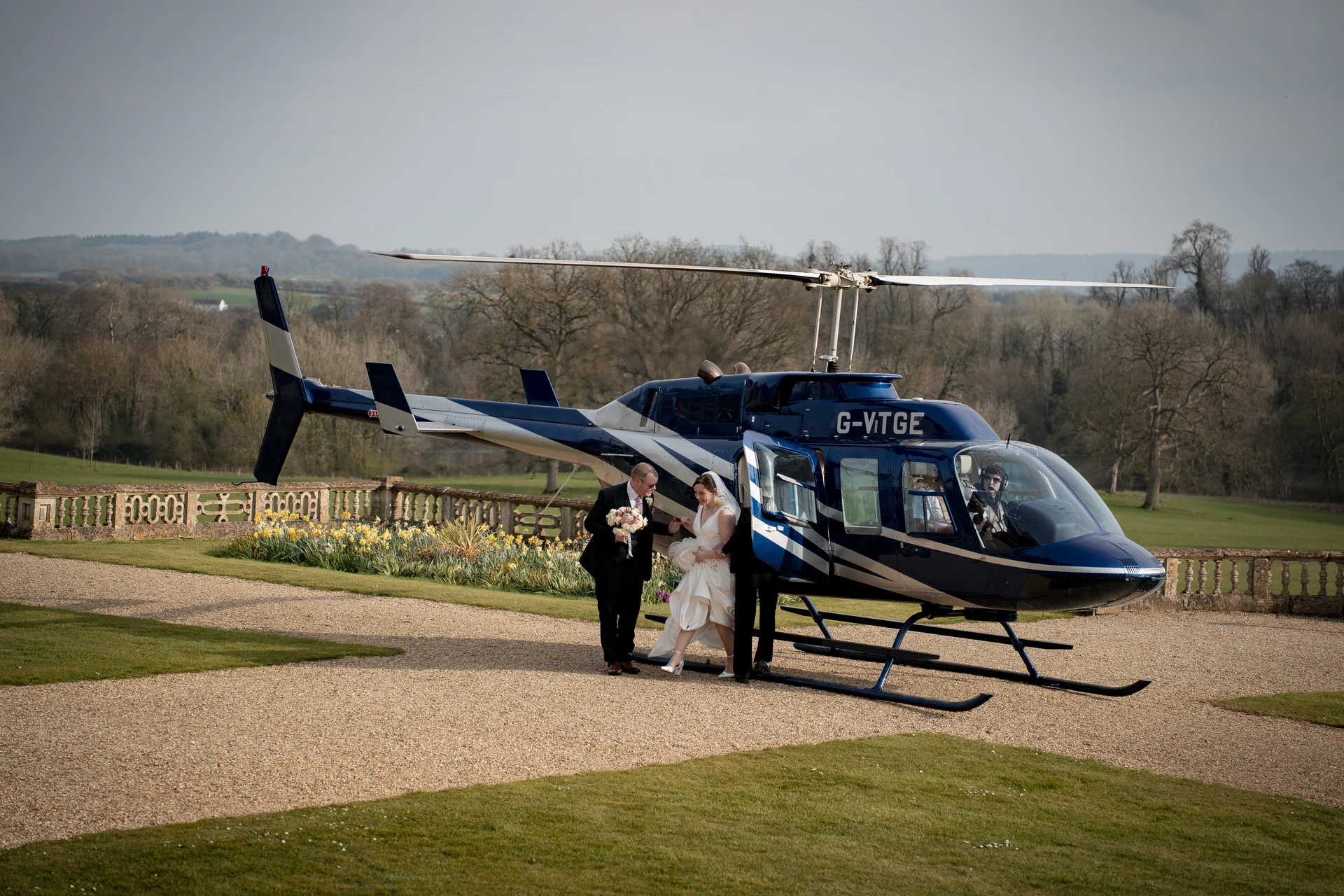 A newlywed couple arrives in style, stepping out of a dark blue helicopter onto the gravel driveway of a grand country house.