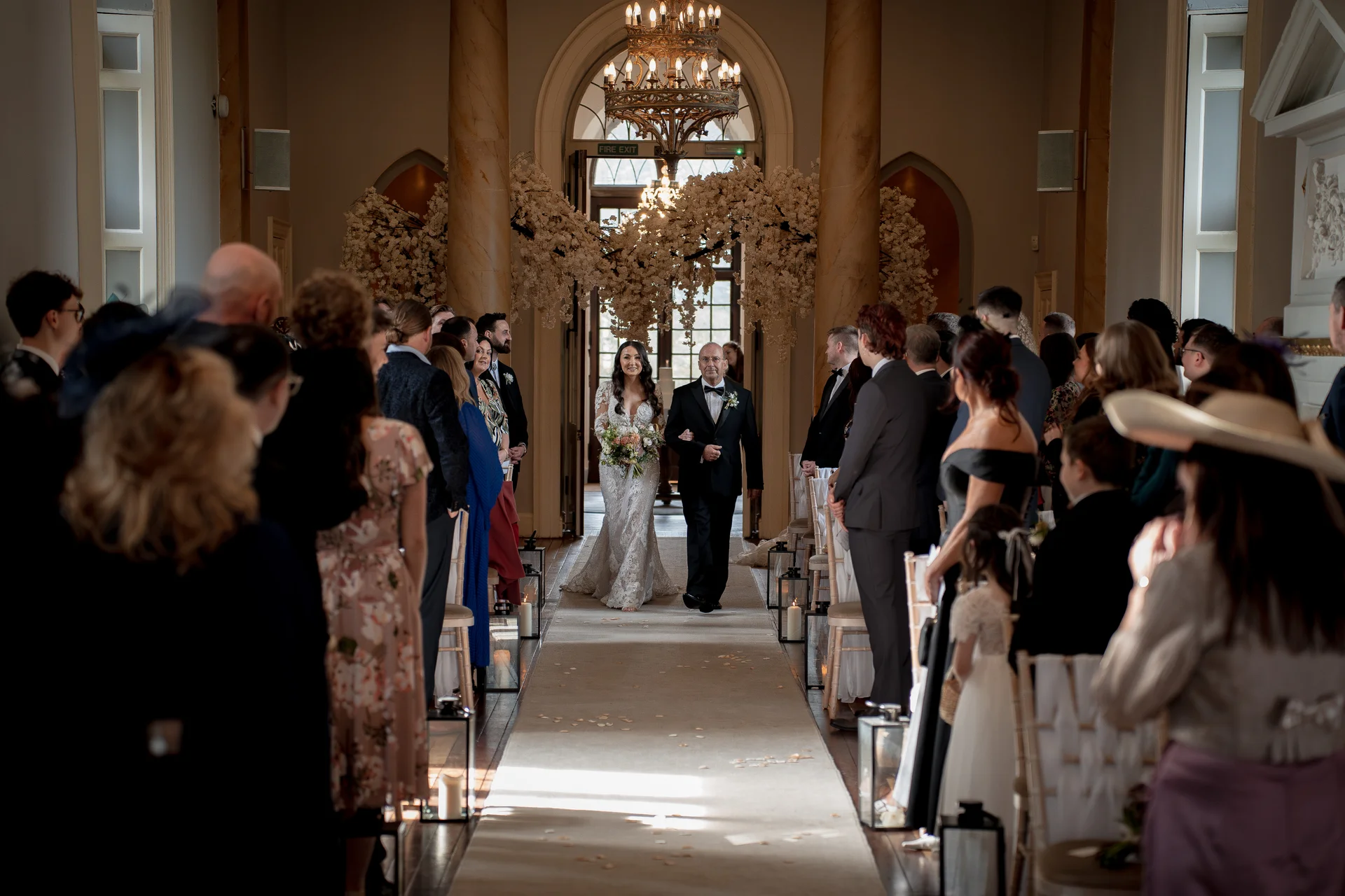 A smiling bride walks down the aisle arm in arm with her father in a grand stately home ceremony room decorated with blossom arches and candlelight.