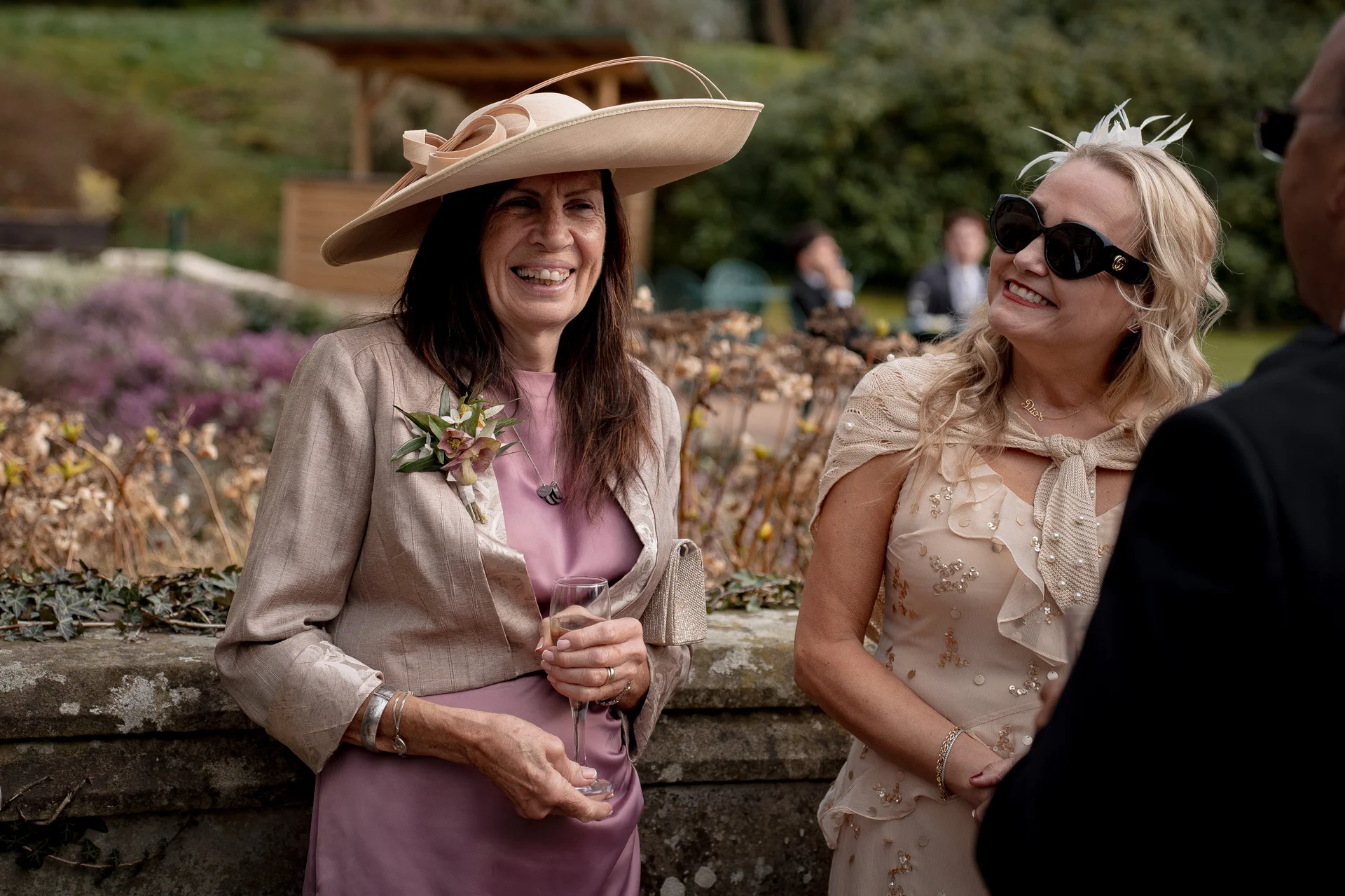 Two elegantly dressed female wedding guests wearing a wide-brim hat and sunglasses chat and laugh together outdoors during a drinks reception.