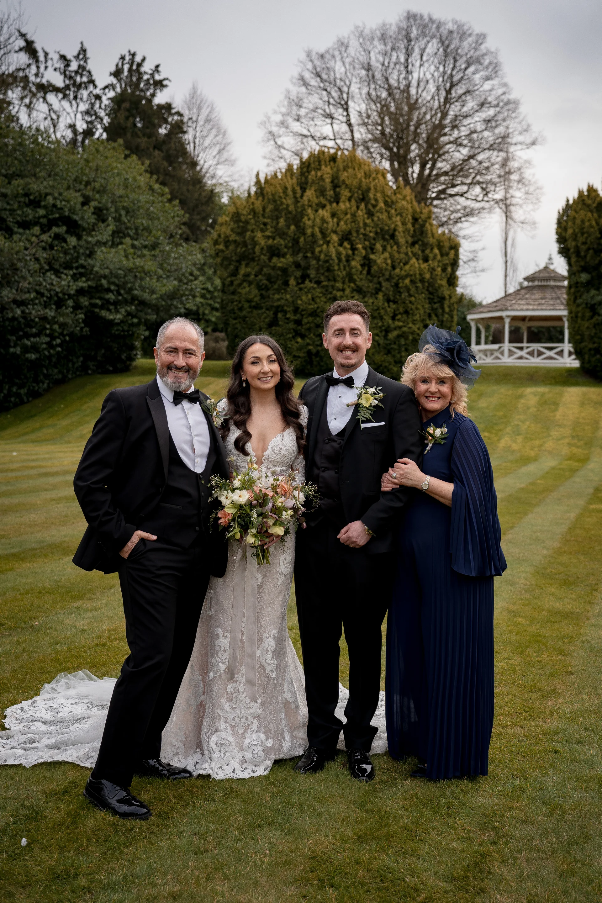 A newlywed couple poses outdoors on a manicured lawn alongside their parents in a formal family wedding portrait.