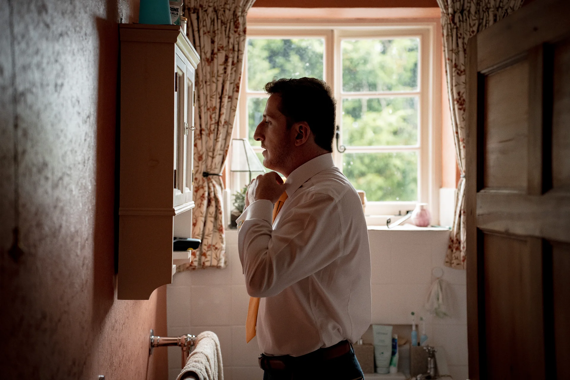 A groom stands in a bathroom getting ready, adjusting his yellow tie while looking into a wall-mounted mirror next to a window.