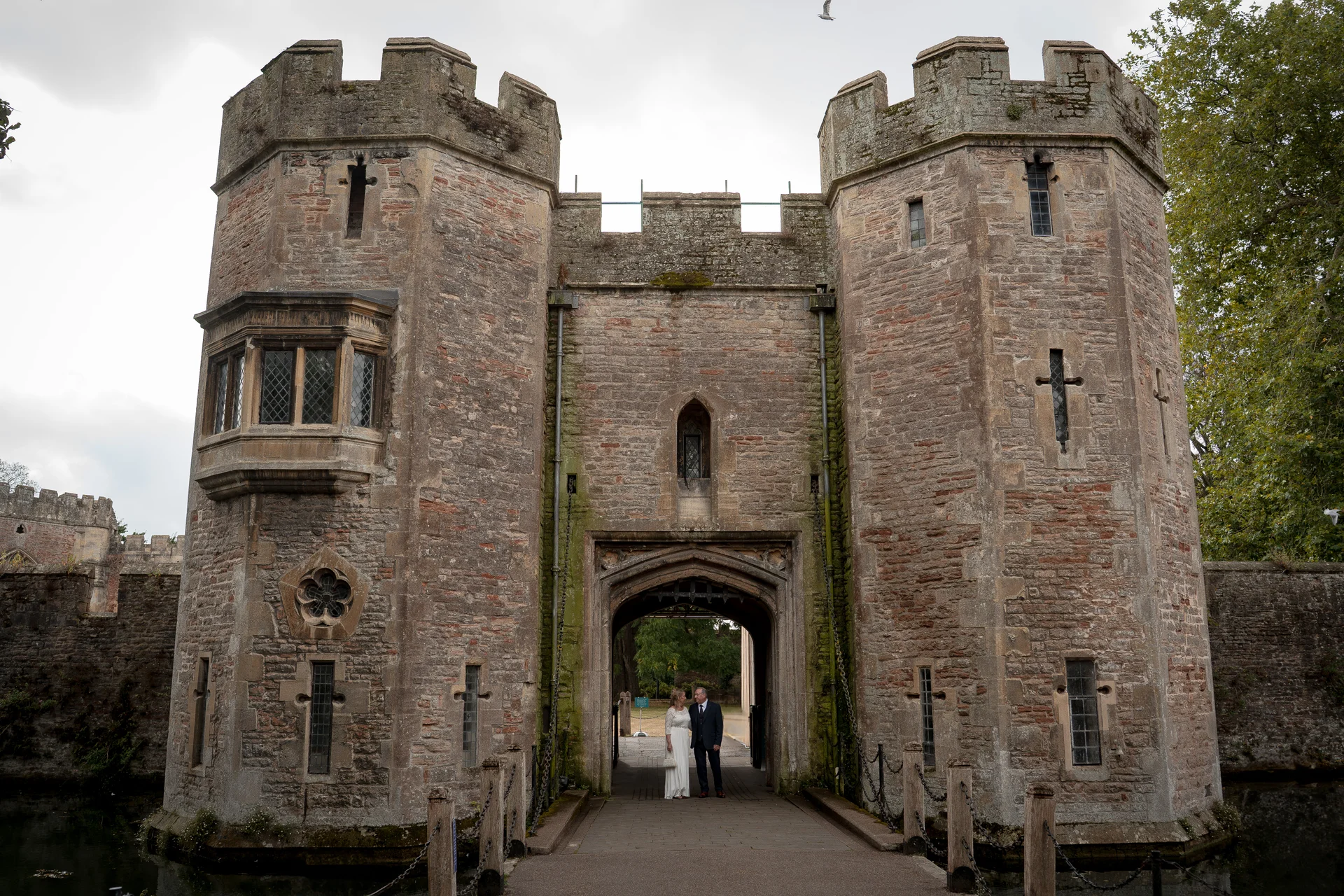 A bride and groom stand in the stone archway of a magnificent medieval castle entrance surrounded by a moat.