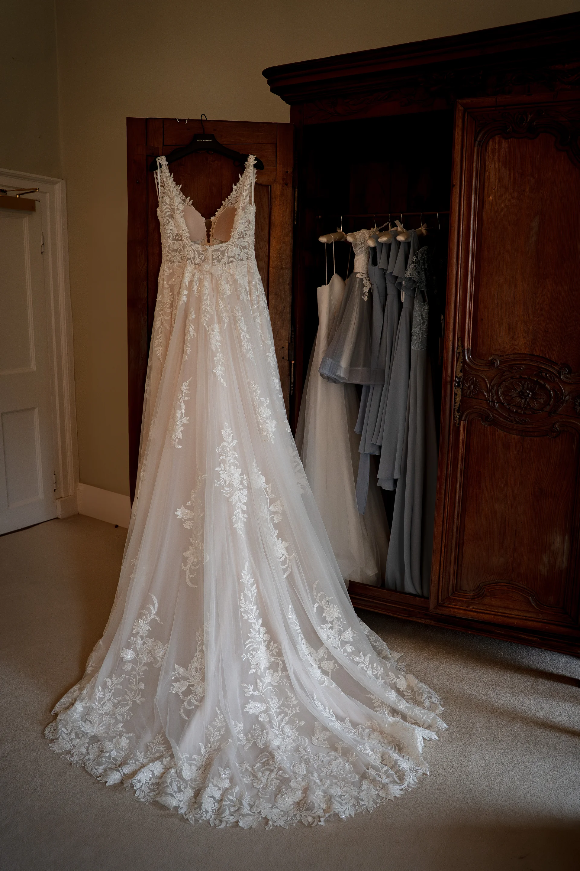 A stunning lace and tulle wedding dress hangs prominently in front of a rustic wooden armoire, with bridesmaid dresses visible inside.