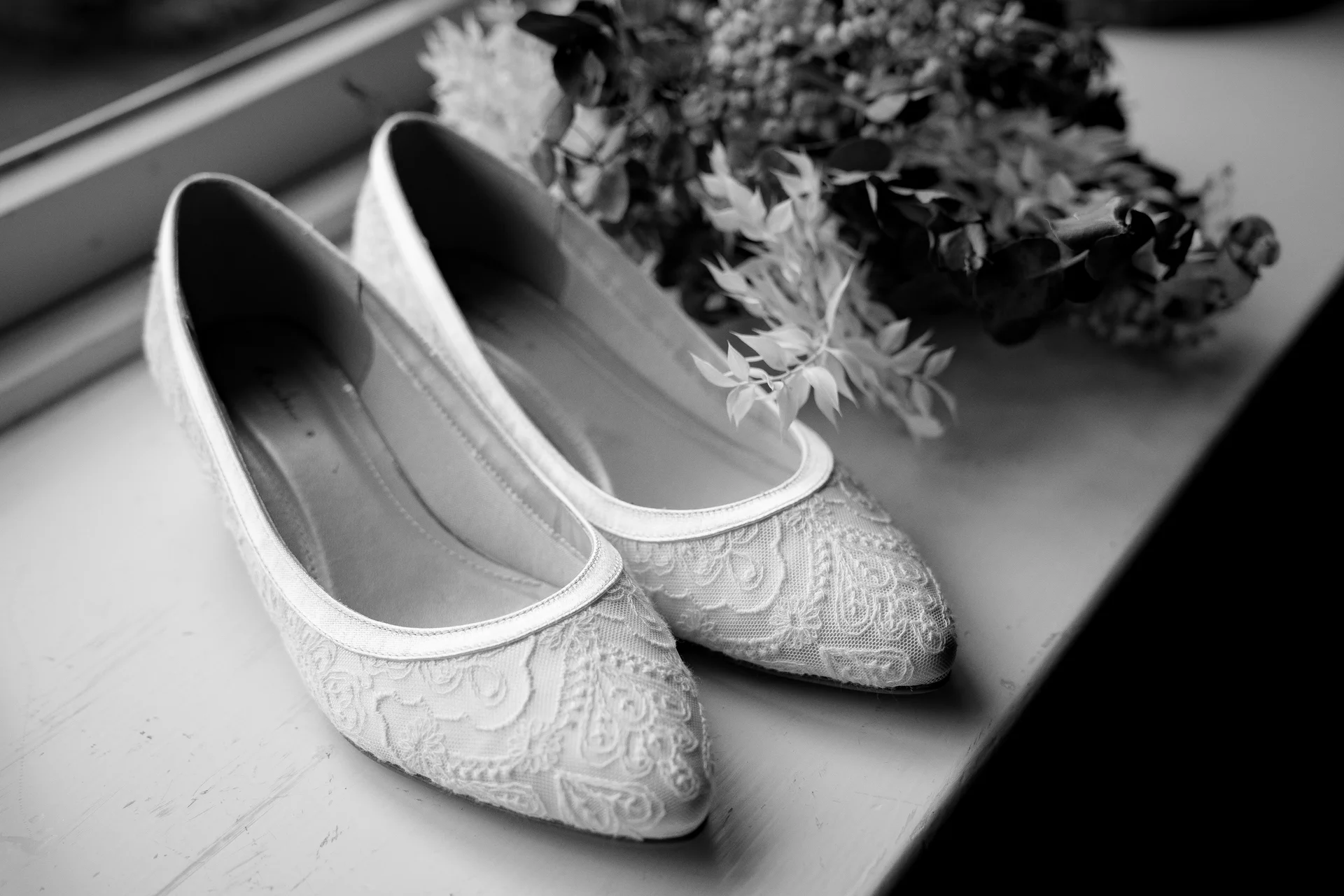 A close-up black and white shot of lace bridal flats resting beside a detailed floral bouquet on a windowsill.