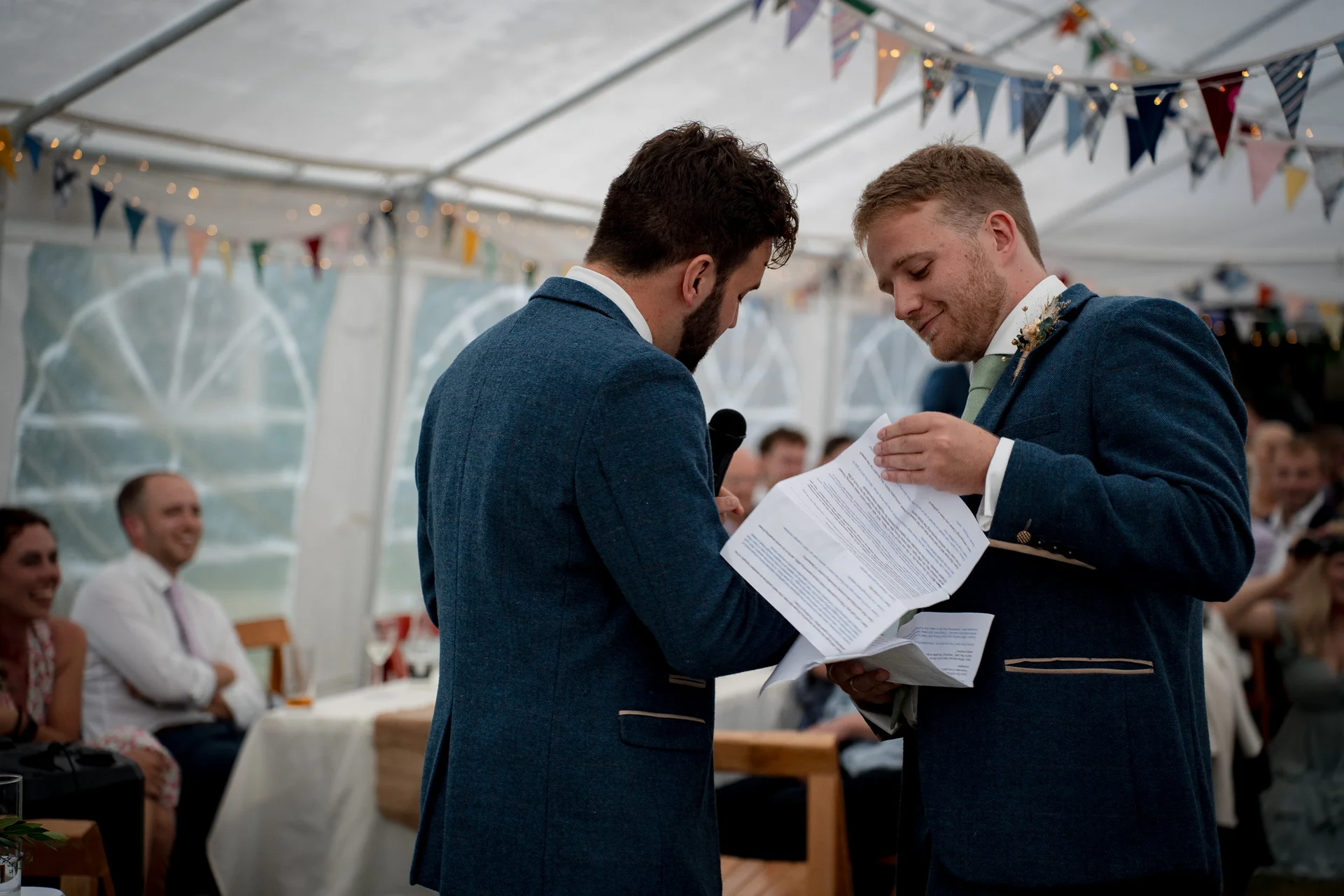 Two grooms in matching tweed suits deliver their wedding speeches inside a marquee decorated with colourful bunting while guests look on and smile.