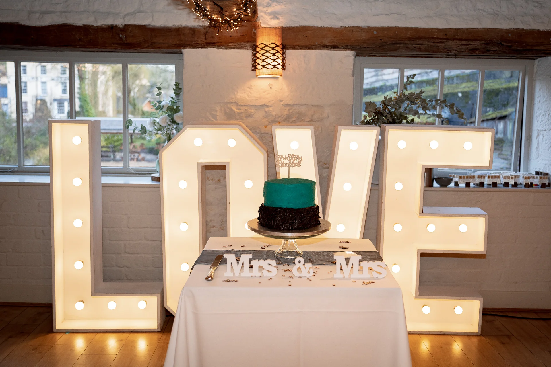 A two-tiered teal and chocolate wedding cake rests on a draped table in front of large illuminated LOVE letters inside a rustic stone barn.