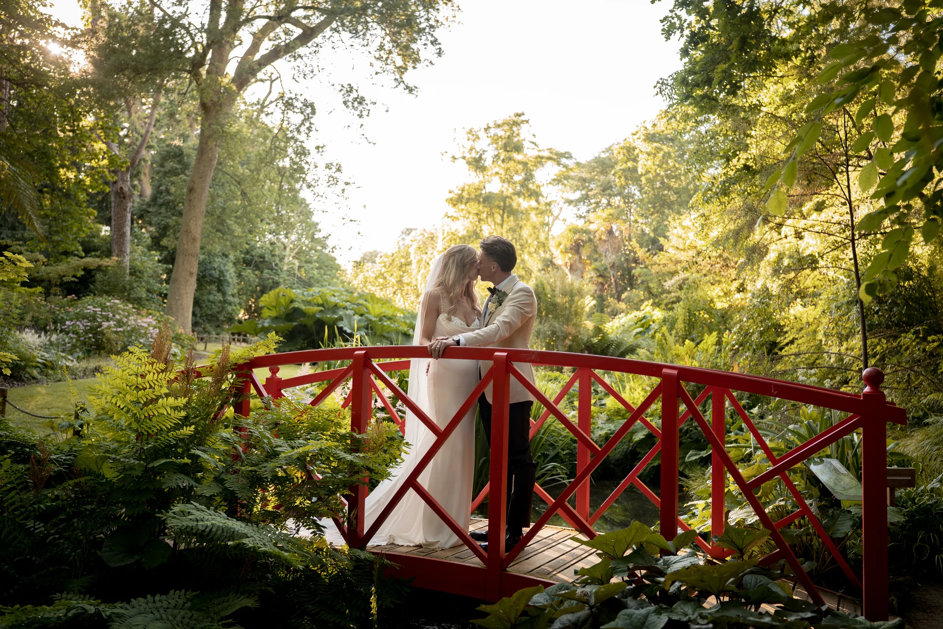 Newlyweds share a romantic kiss on a vibrant red bridge nestled within a lush, sunlit garden during golden hour.