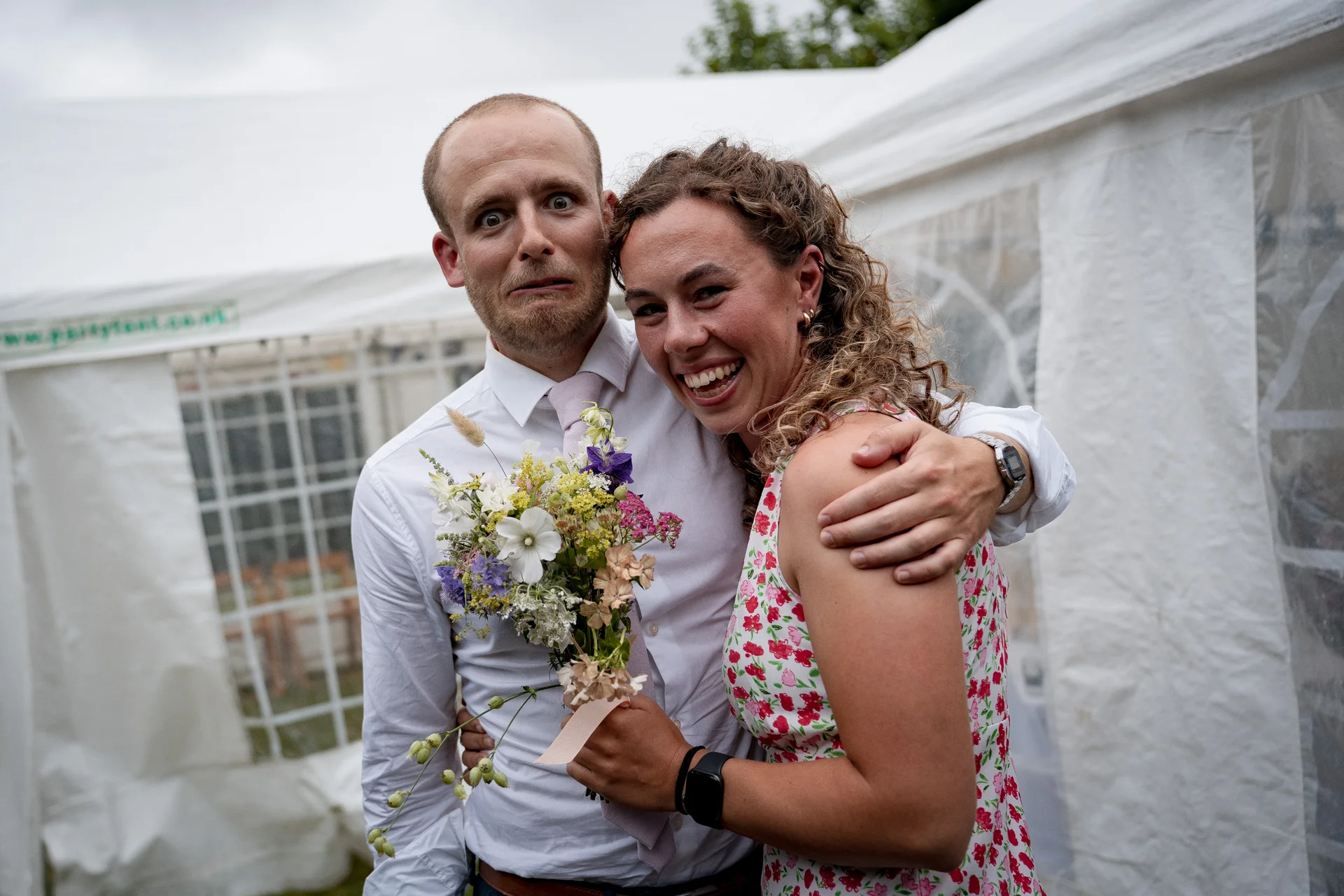 A newlywed couple share a joyful, playful embrace outside a white marquee tent while holding a colourful wildflower bouquet.