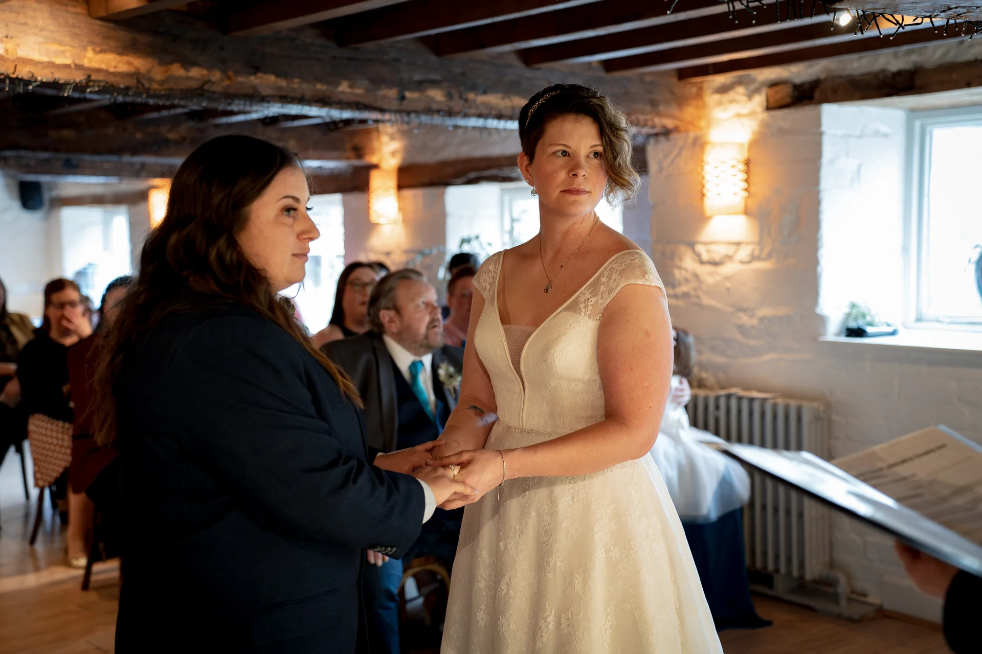 Two brides stand hand in hand during their wedding ceremony inside a rustic barn venue with exposed wooden rafters.