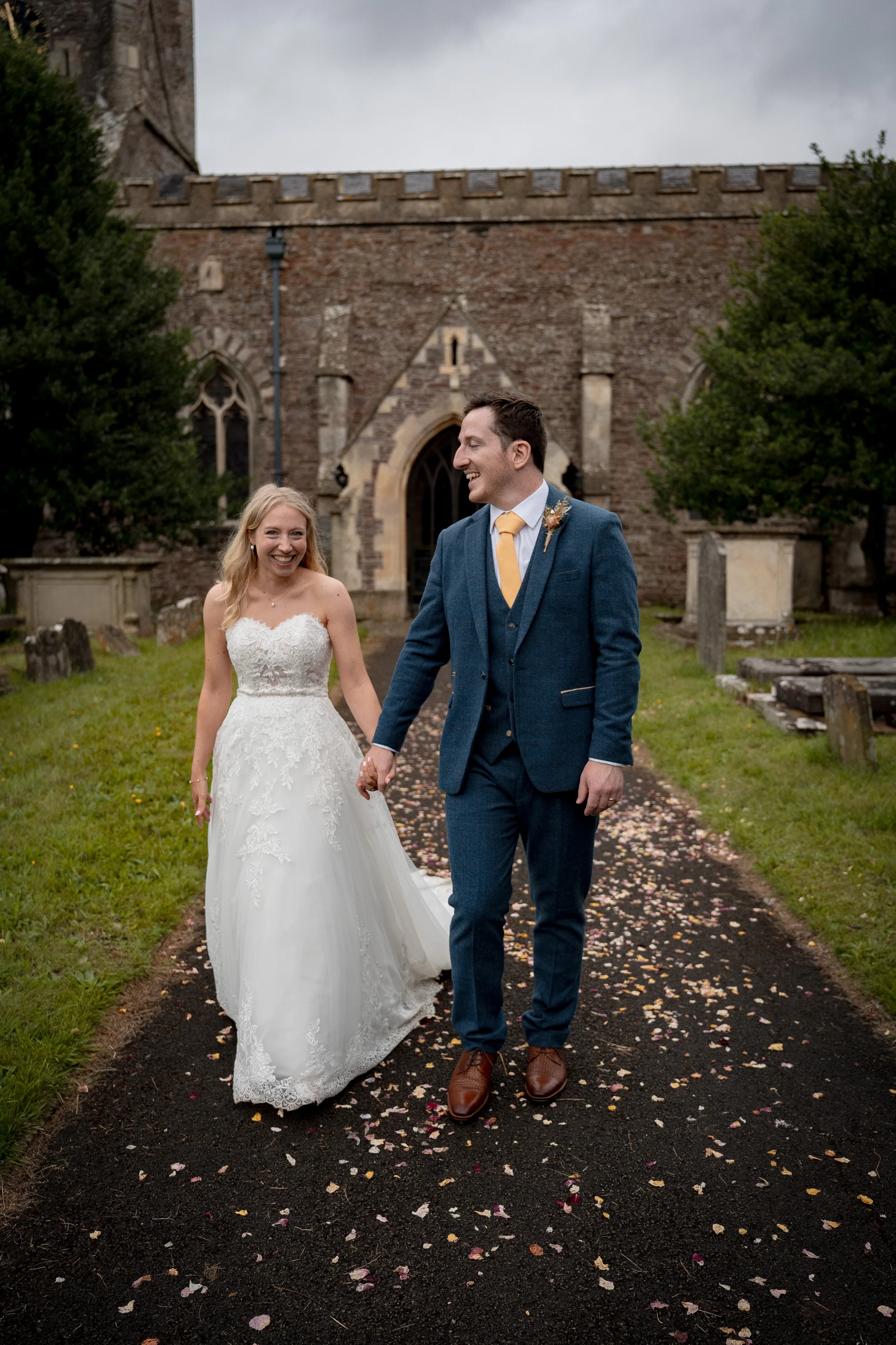 A smiling bride in a strapless lace gown and a groom in a blue tweed suit walk hand in hand along a path strewn with confetti outside a historic stone church.