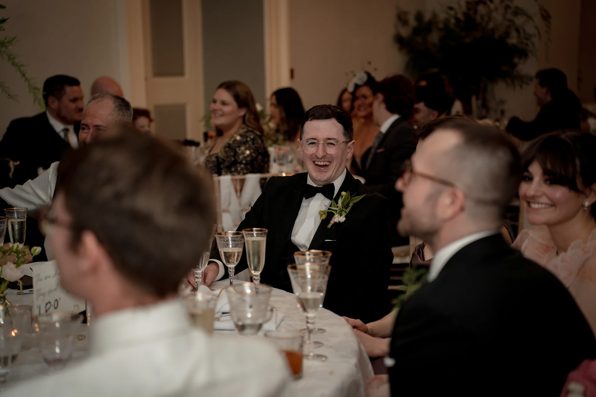 The groom laughing heartily during the wedding speeches while seated at a round table surrounded by smiling guests.