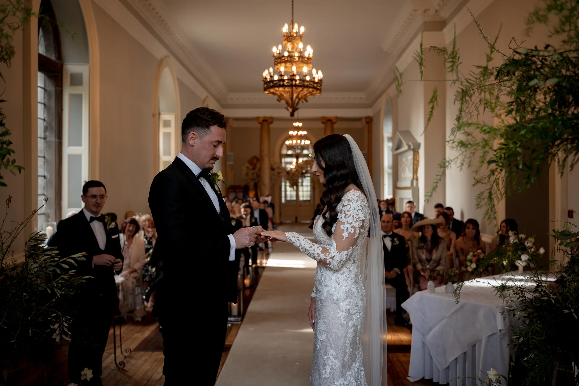 The groom gently places a wedding ring onto the bride's finger during their elegant indoor ceremony in a sunlit historic hall.