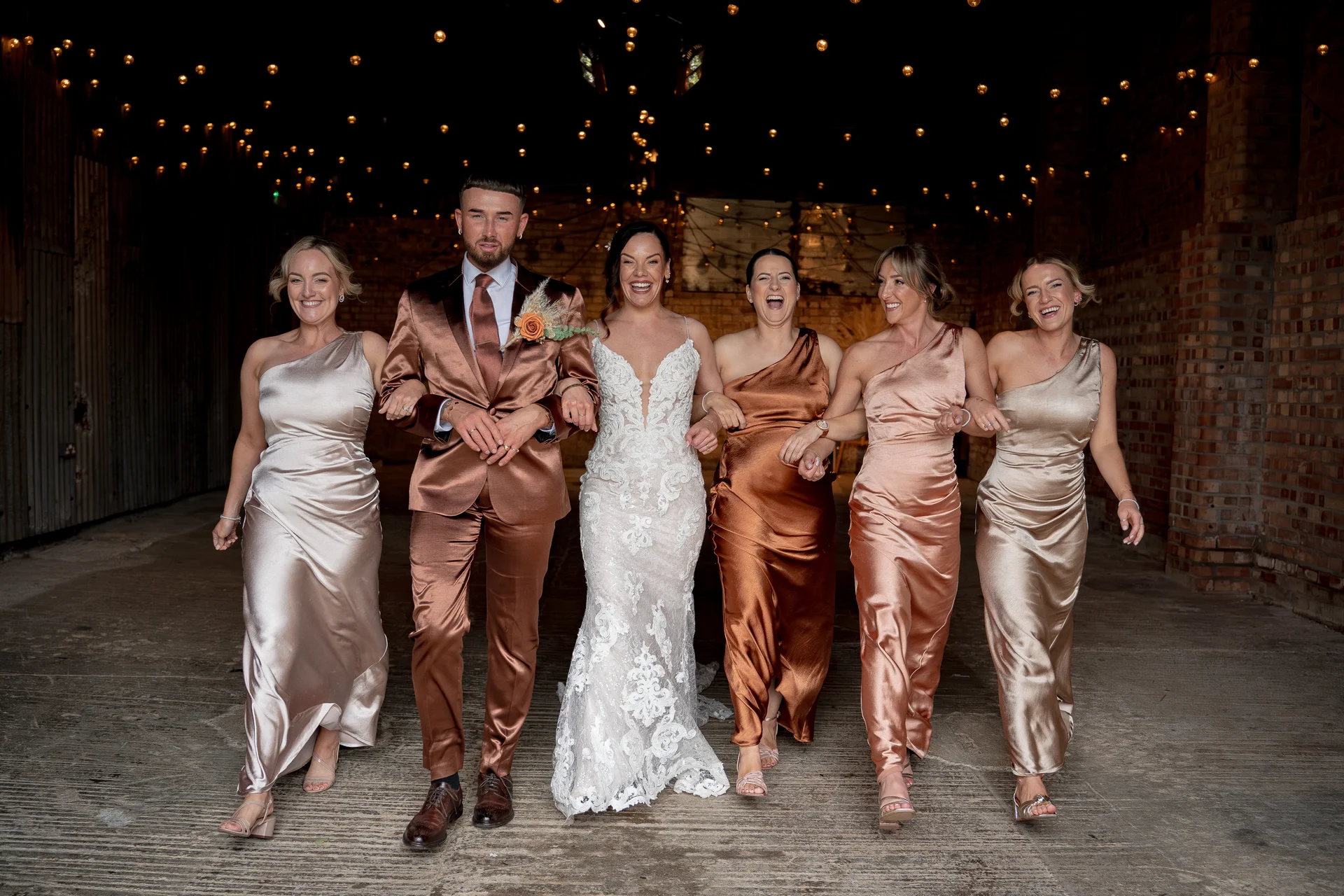 A newlywed couple and their bridal party walk joyfully through a rustic barn illuminated by warm overhead festoon lights.