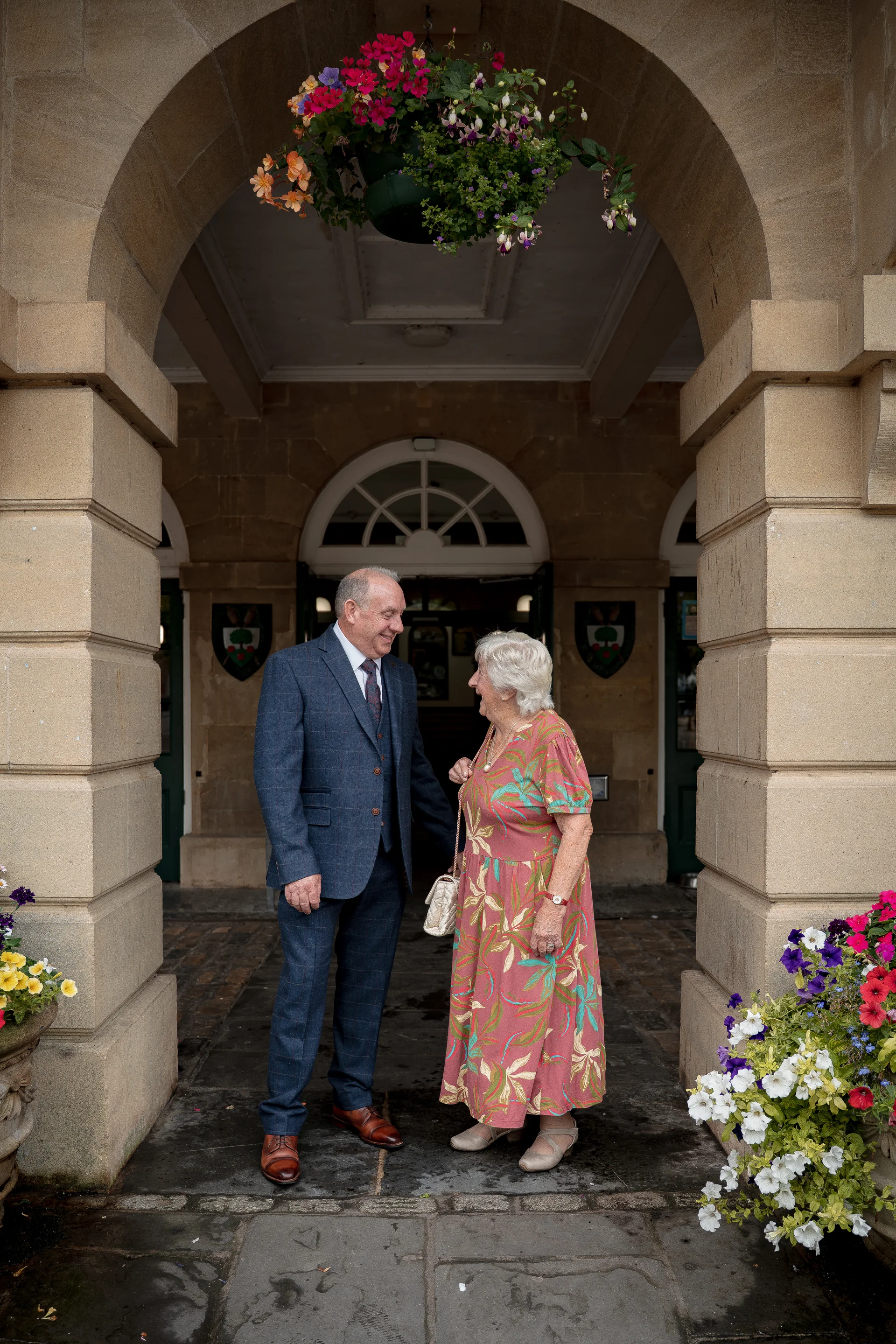 An older couple stand smiling at each other beneath a grand stone archway of a historic venue.