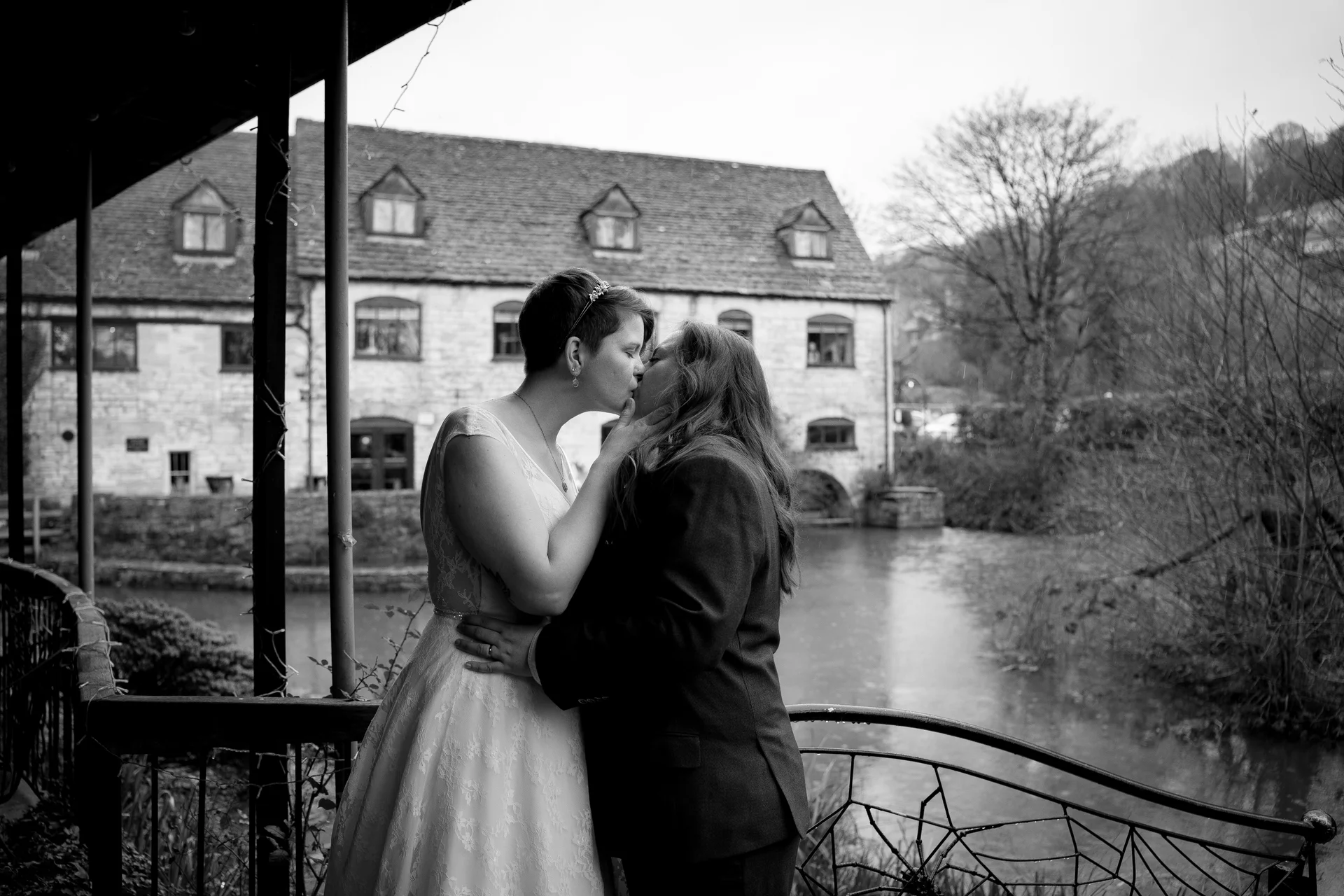 Two brides share a tender kiss on an outdoor terrace overlooking a river and a stone mill on a rainy wedding day.