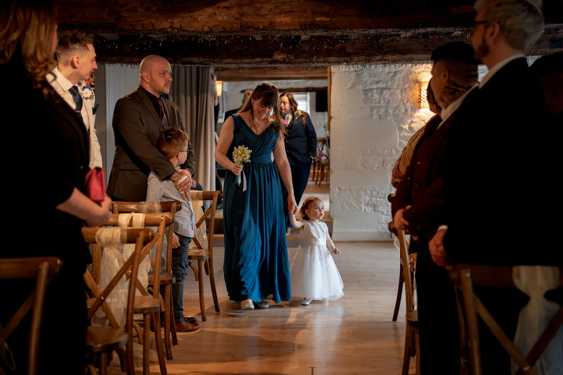 A bridesmaid in a blue dress holding hands with a young flower girl walks down the wooden aisle of a rustic barn wedding ceremony.