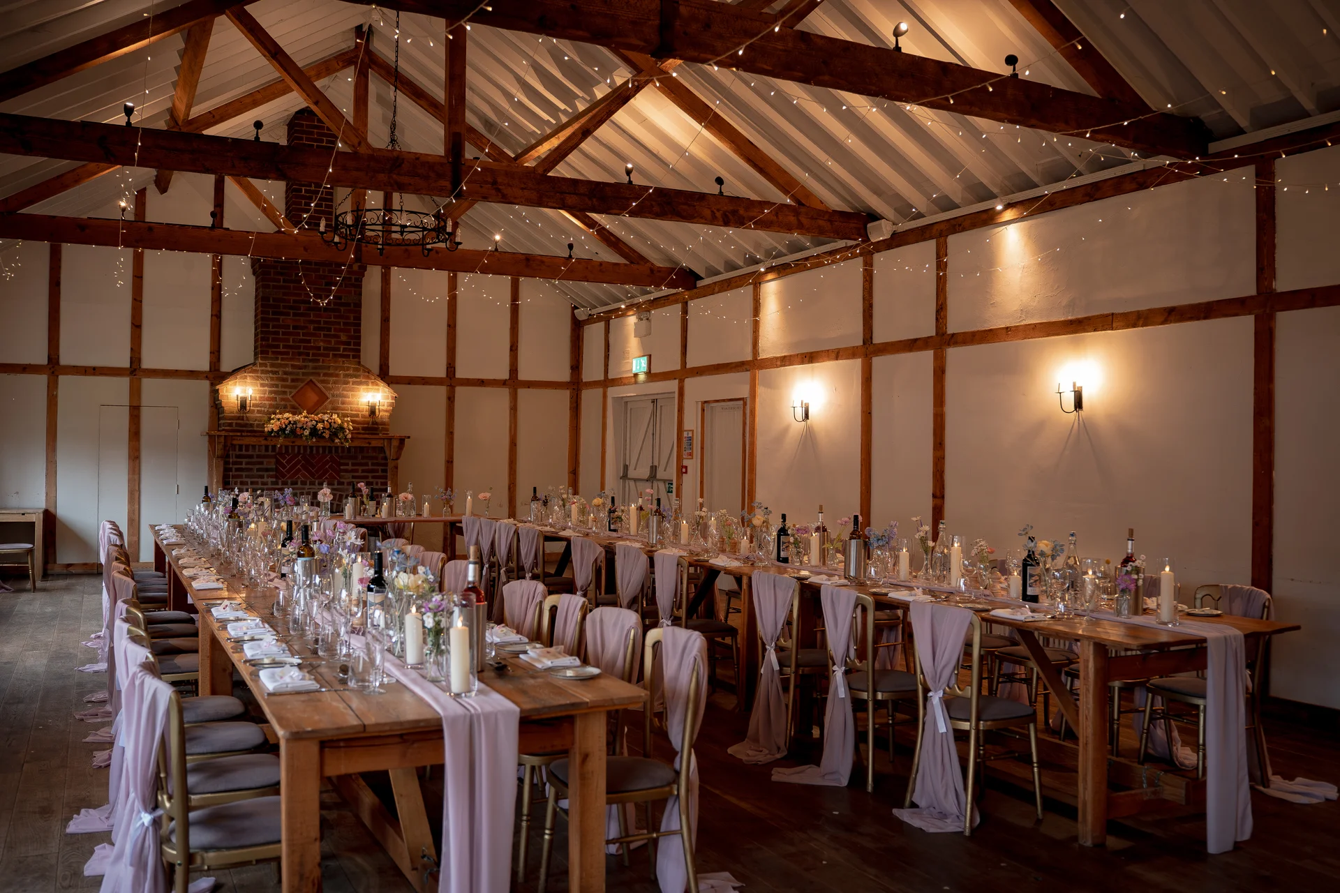 An atmospheric wedding breakfast set inside a rustic oak-framed barn, featuring long wooden banquet tables adorned with dusty rose table runners, tapered candles, and delicate floral arrangements under a canopy of fairy lights.