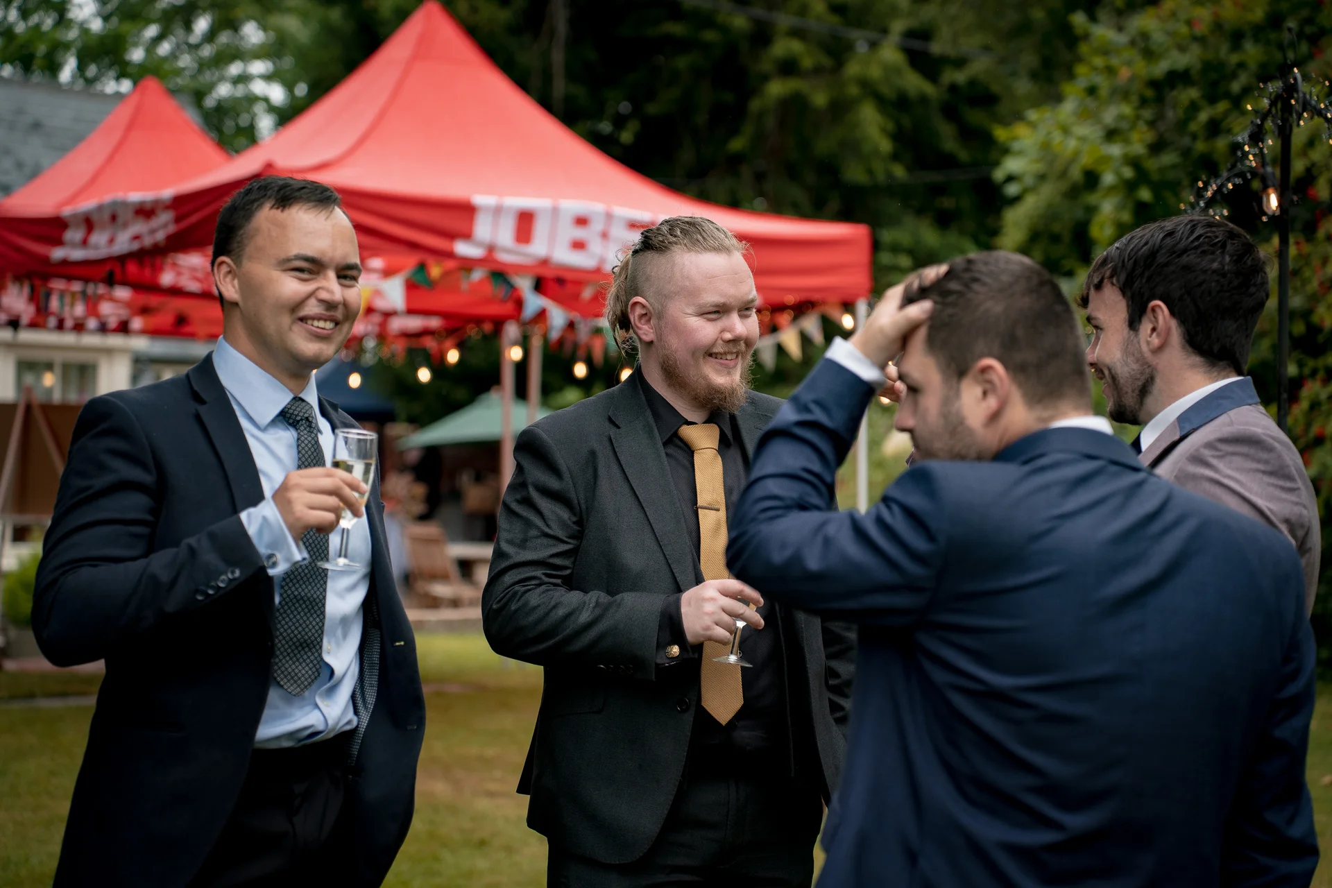 A group of smartly dressed groomsmen and guests mingle and enjoy drinks outside near a red marquee during a wedding celebration.