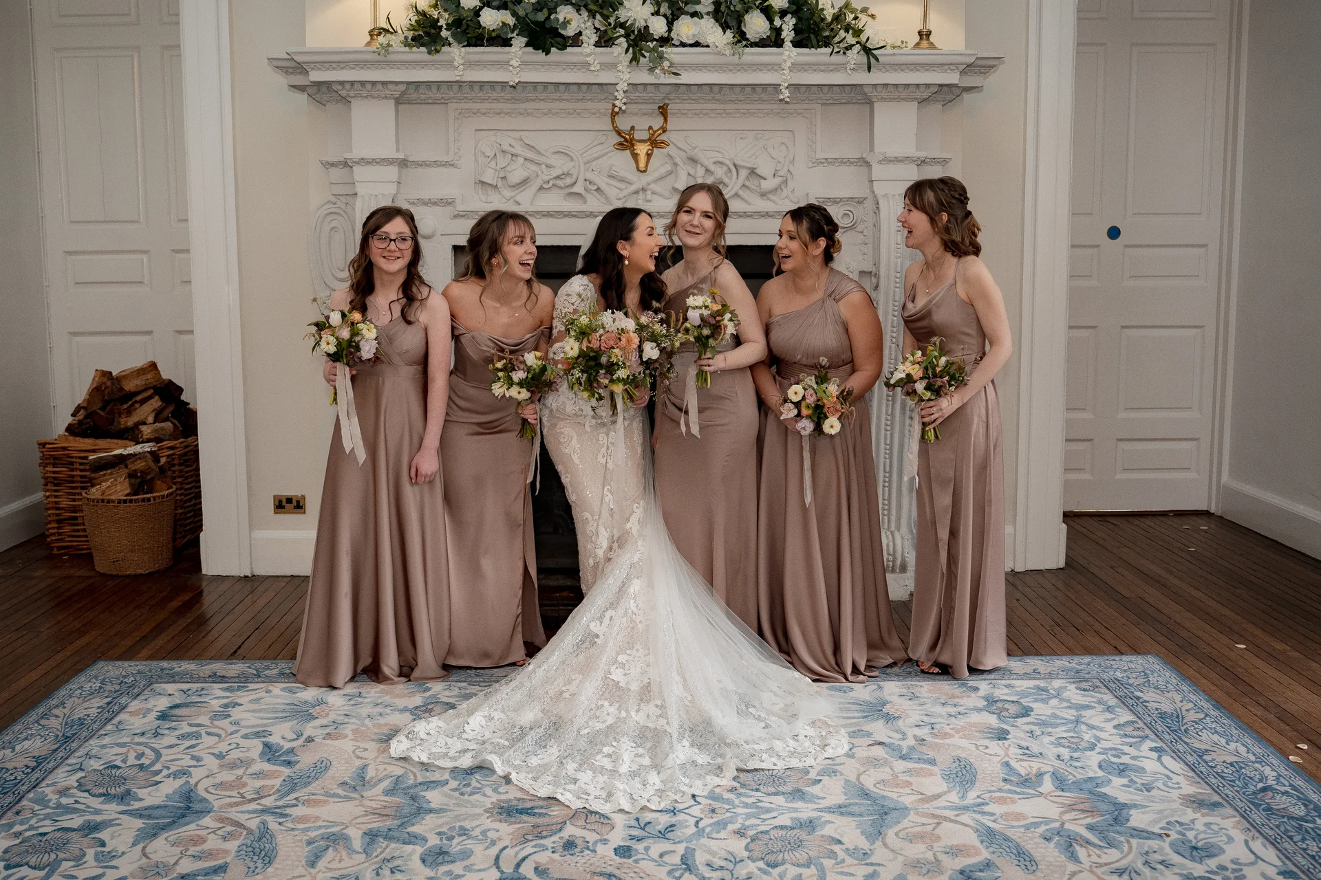 A glowing bride in an intricate lace gown with a dramatic train stands laughing alongside her bridesmaids in matching satin champagne dresses in front of an ornate fireplace.