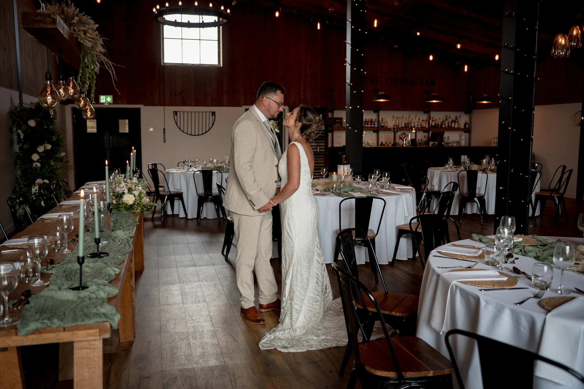 A newlywed couple stands affectionately face to face inside a rustic barn wedding reception venue decorated with long tables and warm candlelight.