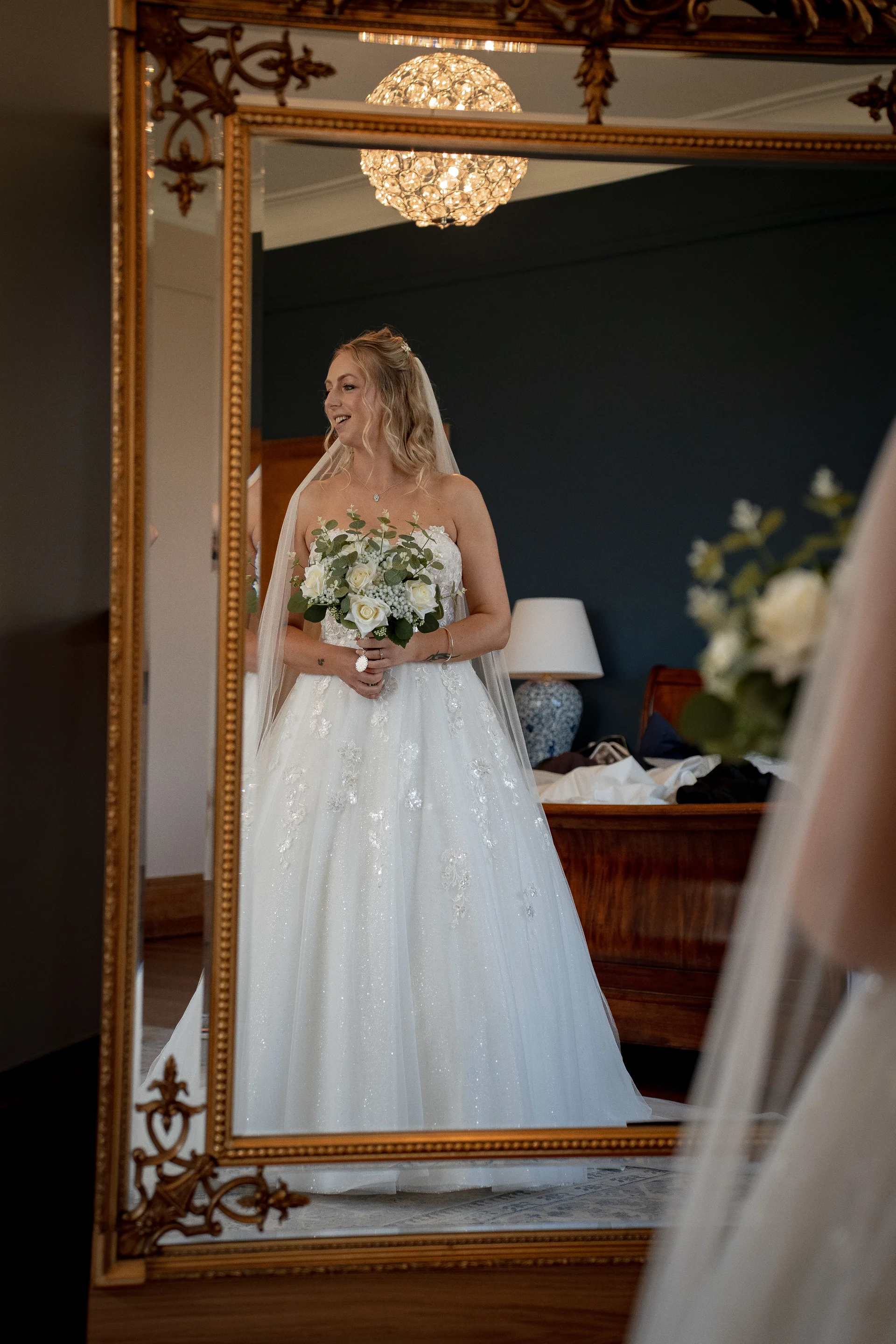 A smiling bride holding a white rose bouquet looks into an ornate gold-framed mirror wearing a strapless a-line gown and veil.