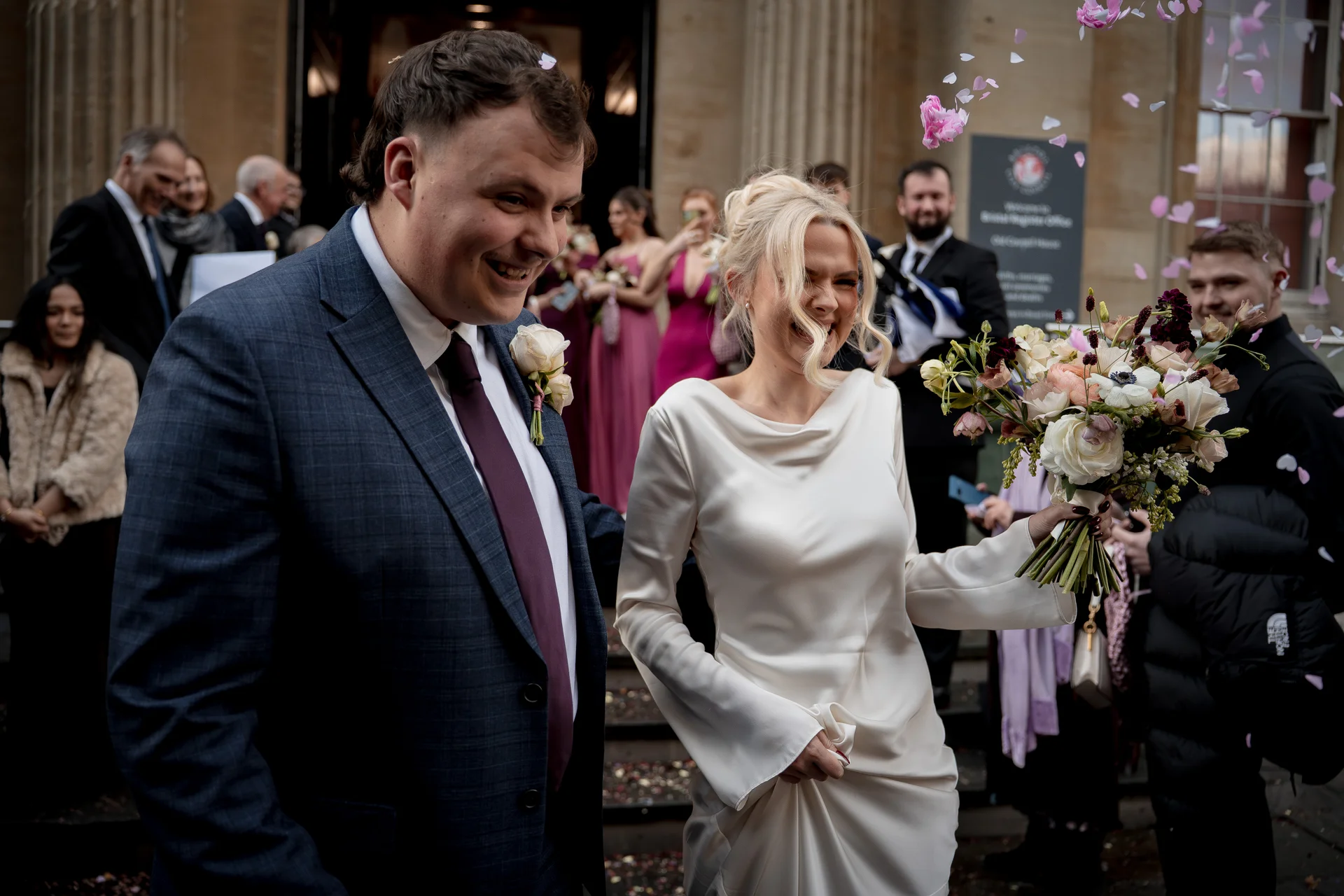 Newlyweds laughing joyfully as confetti falls around them outside a registry office, with the bride holding a lush bouquet and wearing a sleek long-sleeved gown.