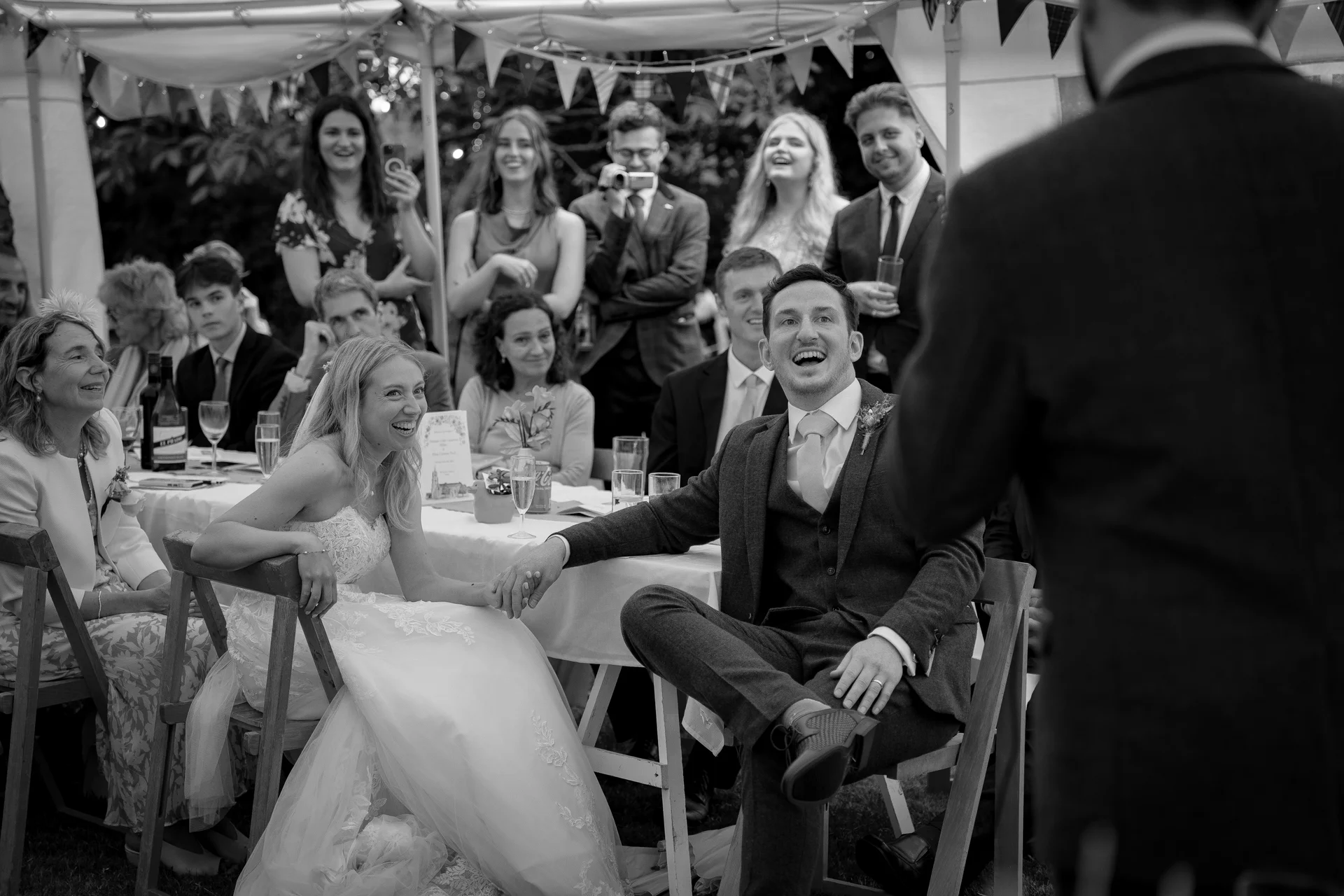 A joyful bride and groom laugh heartily during the wedding speeches inside a marquee while seated at the top table.