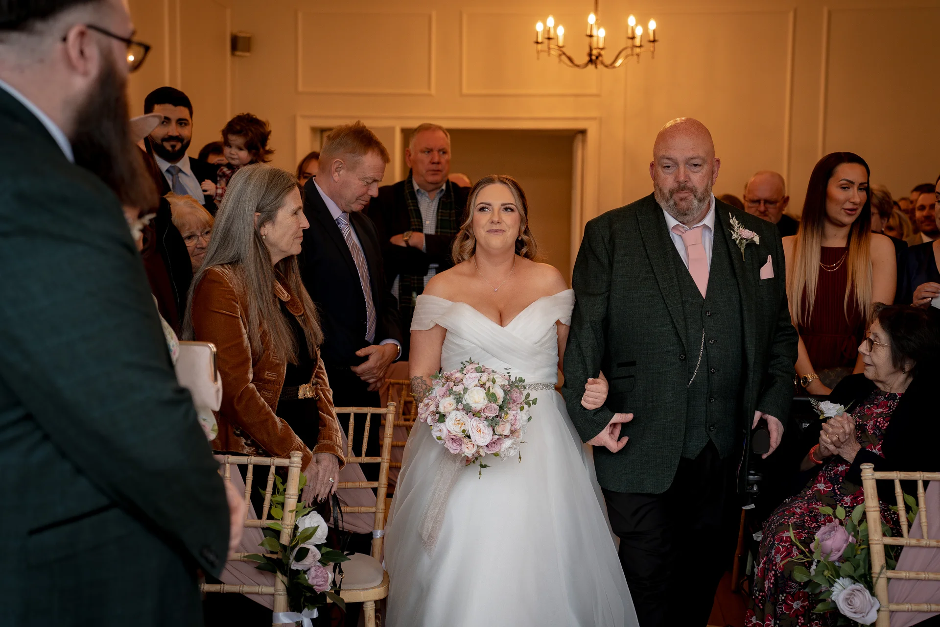 A smiling bride walks down the aisle arm in arm with her father, holding a soft pink and white floral bouquet inside a brightly lit venue.