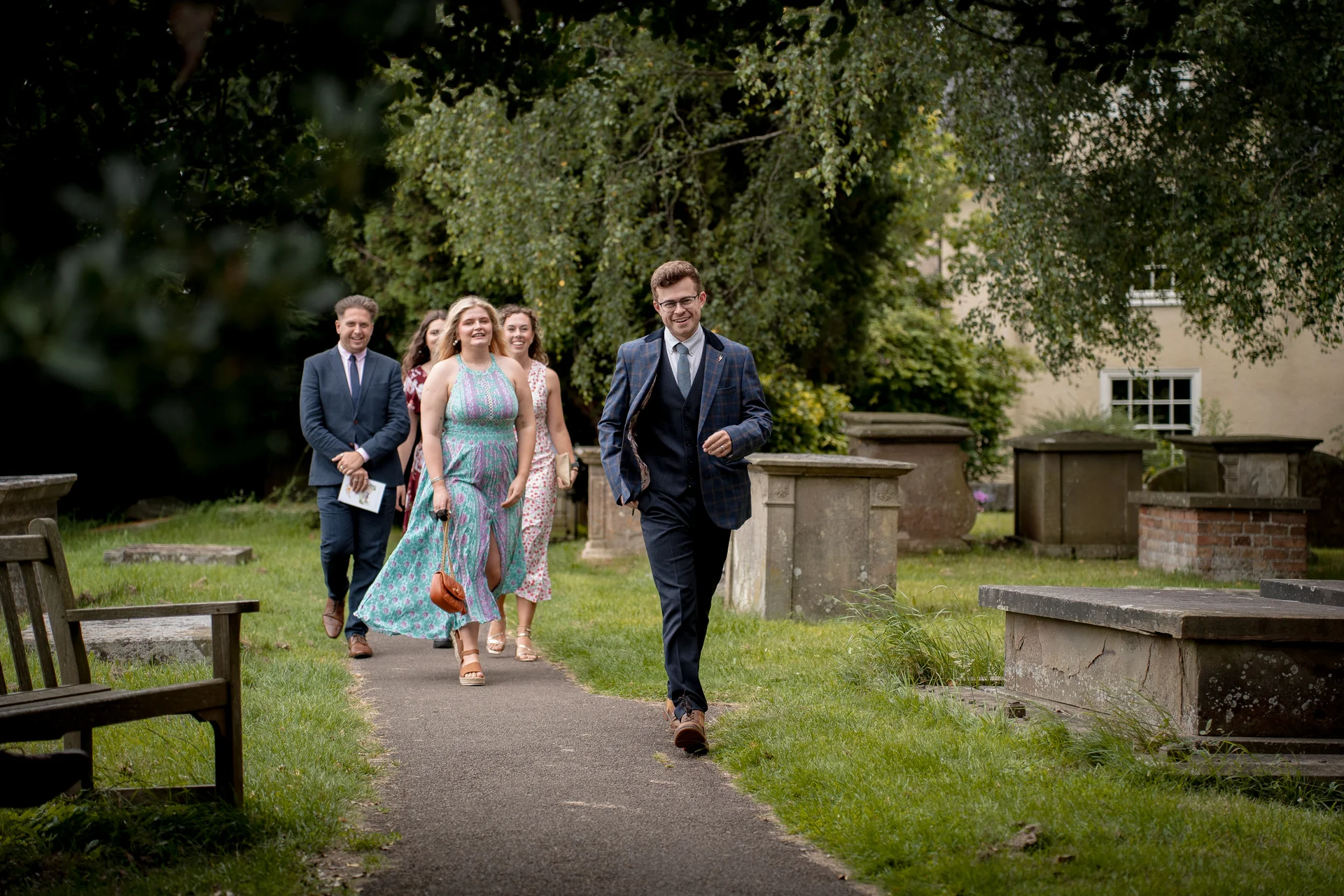 A smiling groom in a checked suit leads a group of wedding guests walking along a path in a lush churchyard surrounded by historic stone tombs and trees.