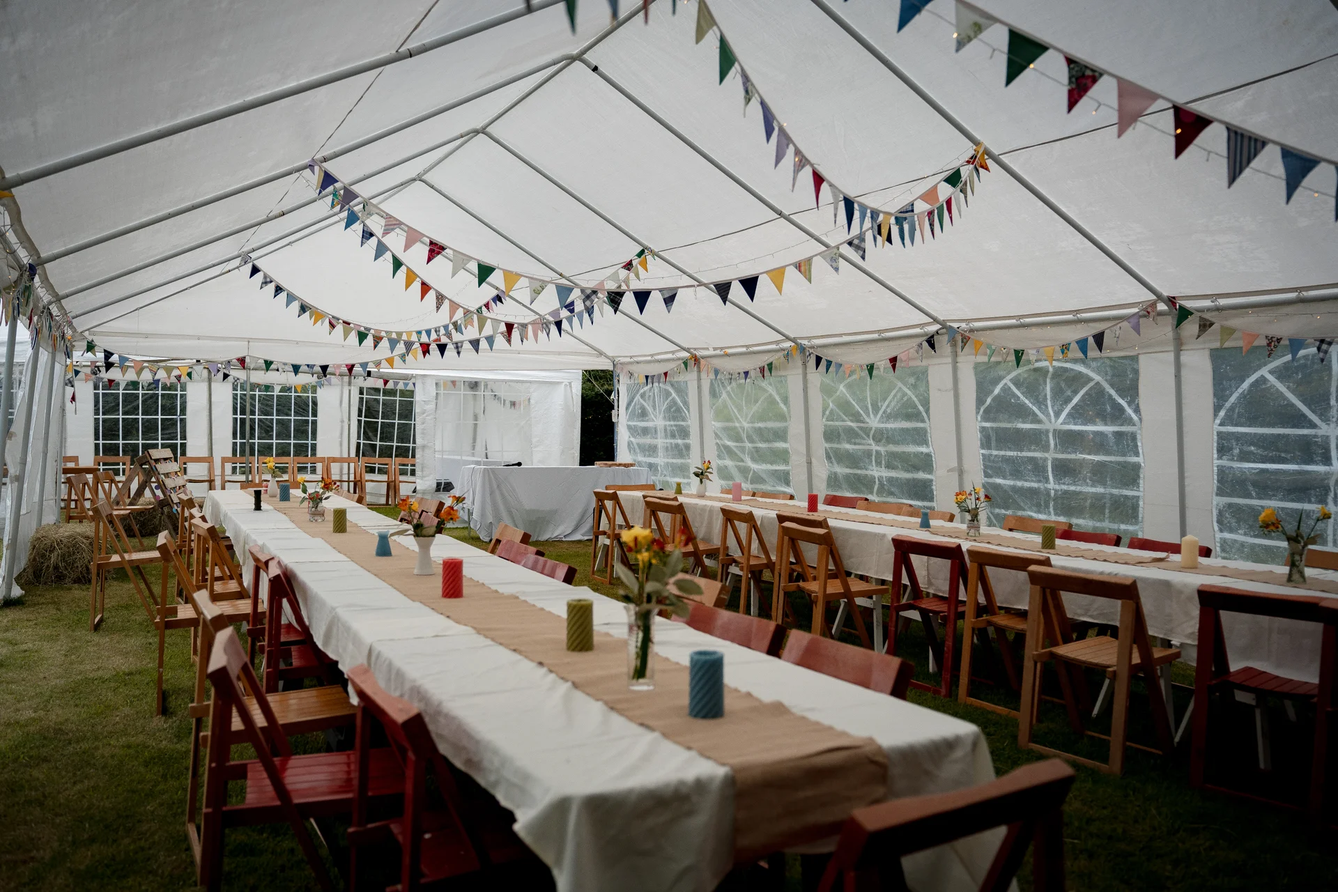 An inviting marquee wedding reception features long banquet tables set up on the grass, decorated with simple runners, colourful pillar candles, and bunting strung across the white ceiling.