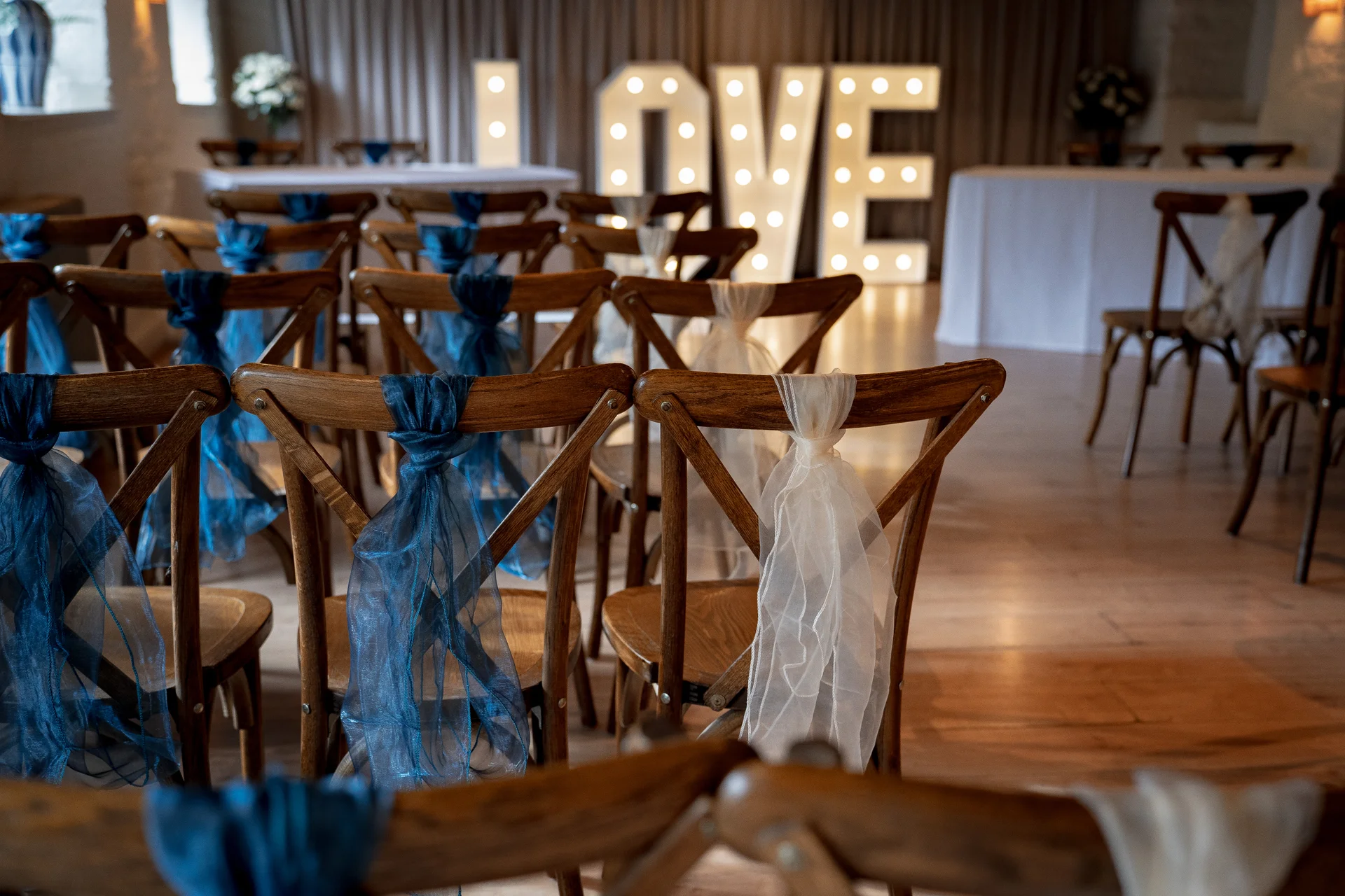 Rows of wooden cross-back chairs are elegantly decorated with flowing blue and white tulle fabric inside a rustic wedding reception venue.