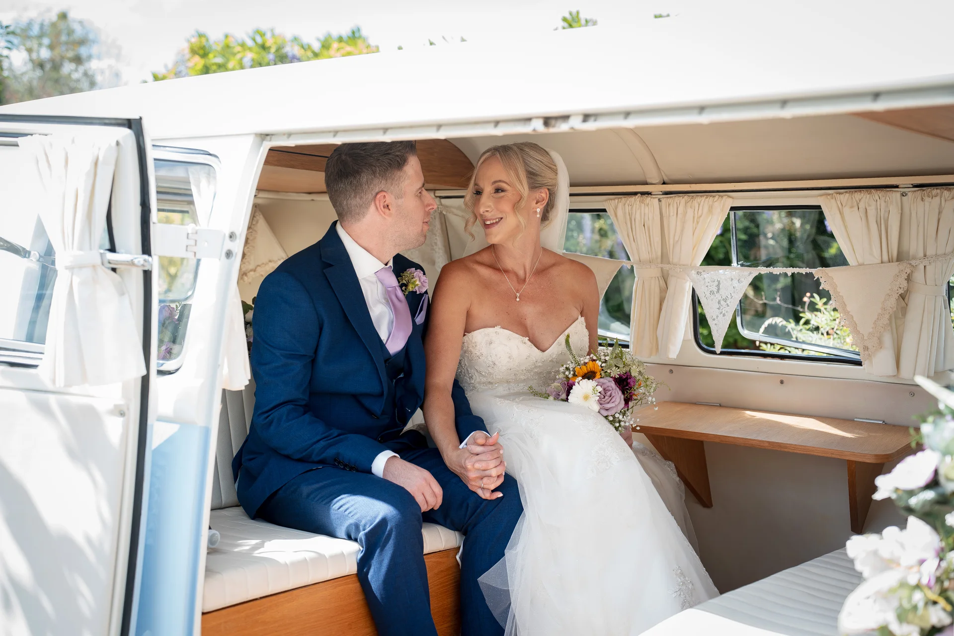 A newlywed bride and groom smiling at each other while holding hands inside the back of a vintage VW campervan getaway car.
